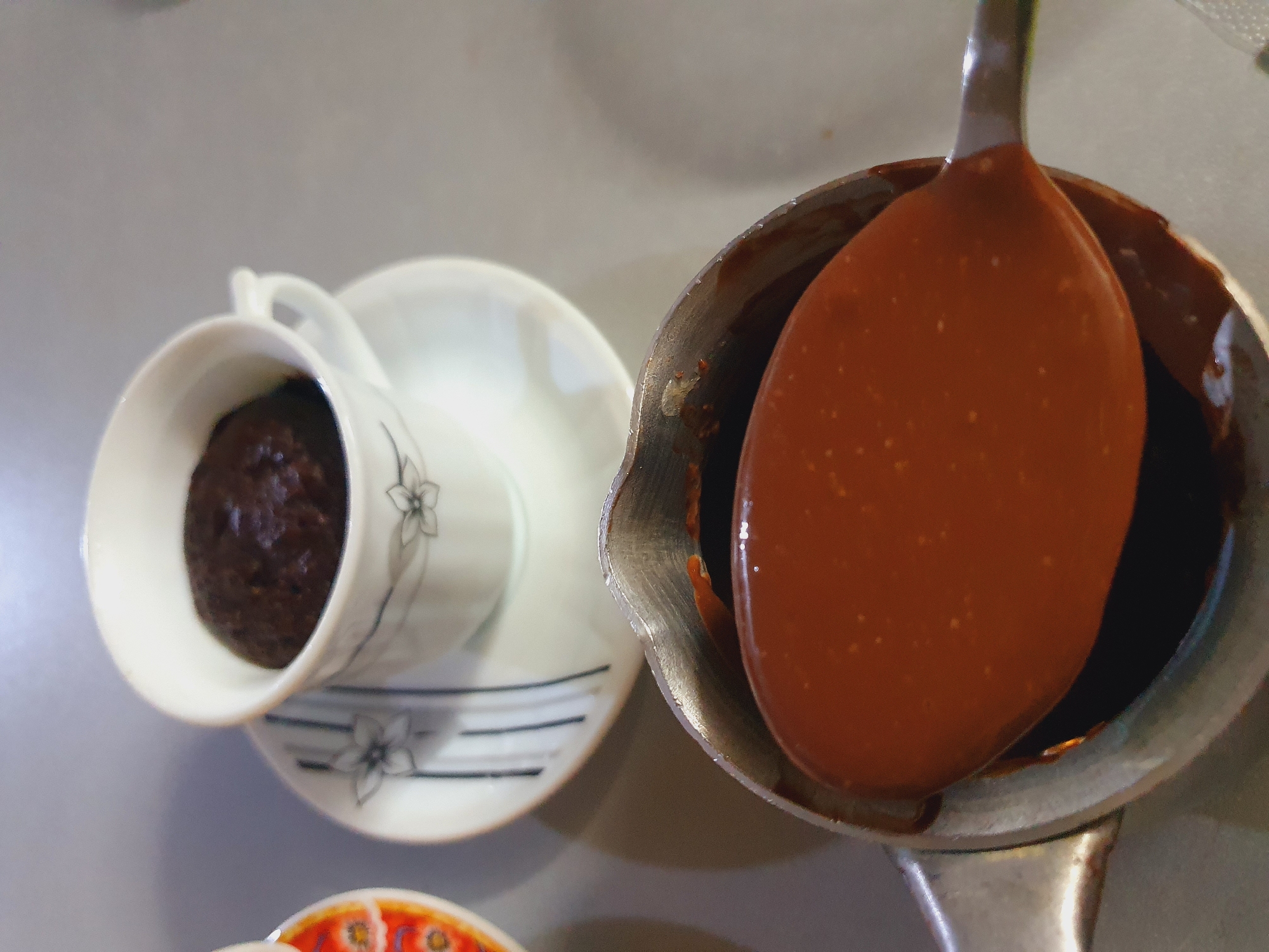 A close-up shot shows a spoon being lifted from a metal pot containing a dark, viscous liquid, likely molten chocolate. The spoon is covered in the liquid, with drips falling back into the pot. In the background, a white teacup sits on a saucer, containing a dark, dense food item, possibly a small cake or a portion of a dessert. The teacup is decorated with a floral pattern and vertical lines. To the left of the teacup, a sliver of another dish with an orange and floral pattern is visible. The surface the objects are resting on appears to be a light gray countertop. The image is taken from a high angle, focusing on the spoon and the pot. There is no information available to determine the location of this scene in the city of Shiraz, Iran.