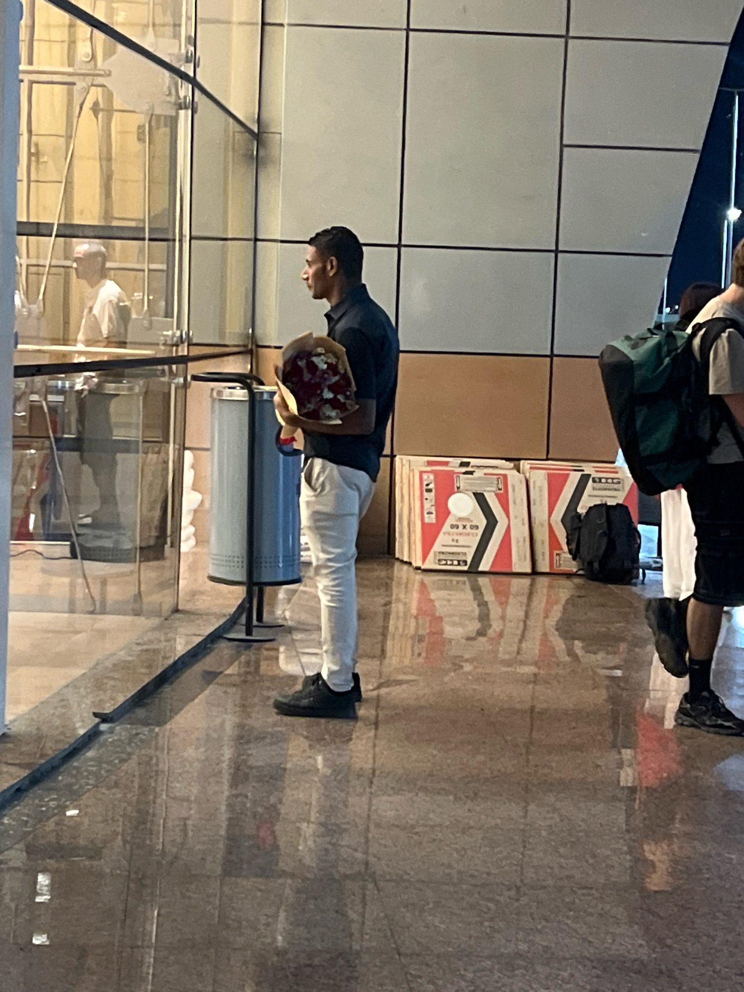 In Sharm El Sheikh, Egypt, a medium-skinned man stands near a glass enclosure, holding a bouquet of red roses. He is dressed in a dark blue collared shirt and light-colored pants, with black sneakers. To his left, a blue trash receptacle is visible. Behind him, several cardboard boxes with orange and white markings are stacked, and a dark backpack rests on the floor. Further to the right, another individual wearing a backpack is partially visible. The environment features a polished floor that reflects the surroundings, suggesting an indoor setting such as an airport terminal or a modern building.