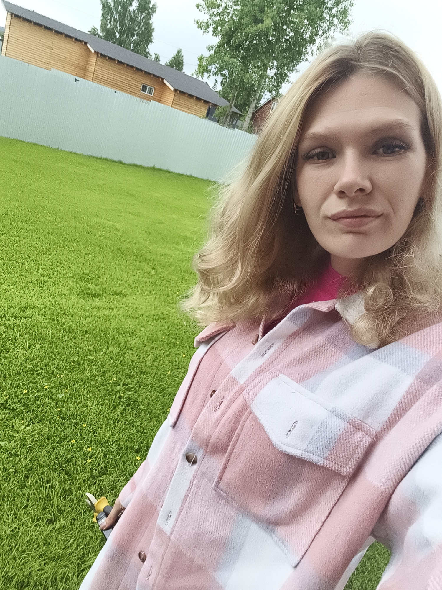 A young woman with blonde hair and makeup poses for a selfie outdoors in what appears to be a suburban or rural setting. She is wearing a pink and white plaid shirt over a bright pink shirt. In her right hand, she is holding a pair of yellow and black garden shears. The background consists of a bright green lawn, a light blue corrugated metal fence, and a wooden building with a dark roof. Trees are visible behind the building and fence. The sky is overcast. The woman is smiling slightly and looking at the camera.