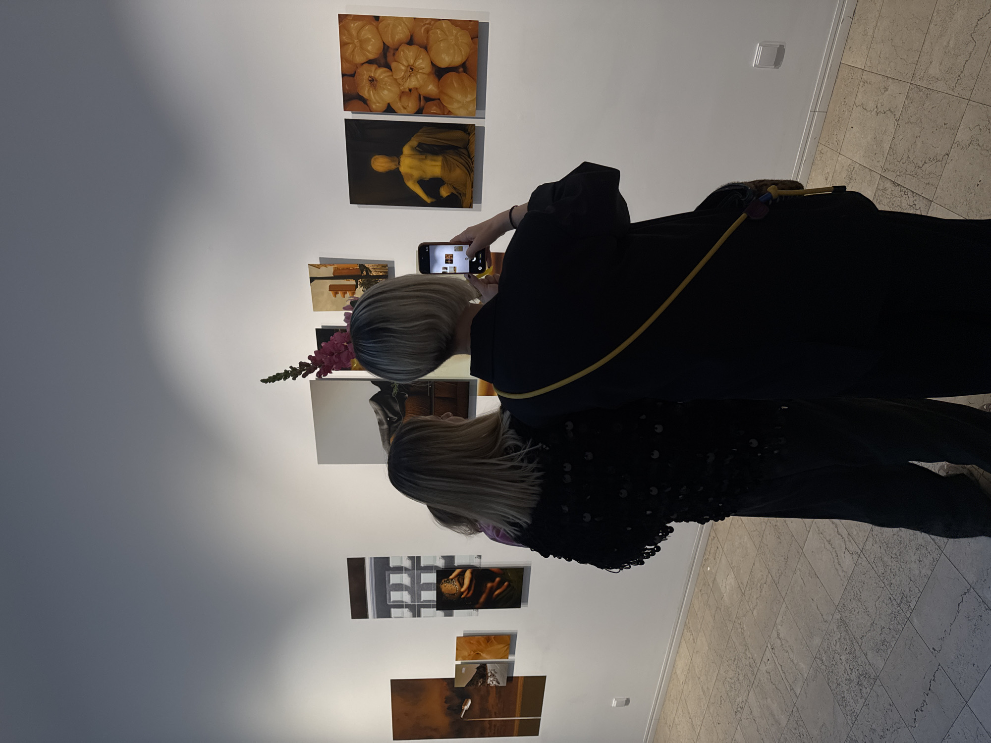 The image captures an art exhibition in Sofia, Bulgaria. Two women are present: one taking a photo with her smartphone and another standing beside her, looking at the artwork. The woman taking the photo has short grey hair and is wearing a black outfit with a yellow stripe. The other woman has long blonde hair and is wearing a black outfit with dark-colored sequins. They are standing in front of a white wall adorned with a series of art pieces. The pieces on display include depictions of fruits, a figure draped in fabric, architectural scenes, and abstract imagery. The room has light-colored tiled flooring, and a light switch is visible on the wall in the upper right.