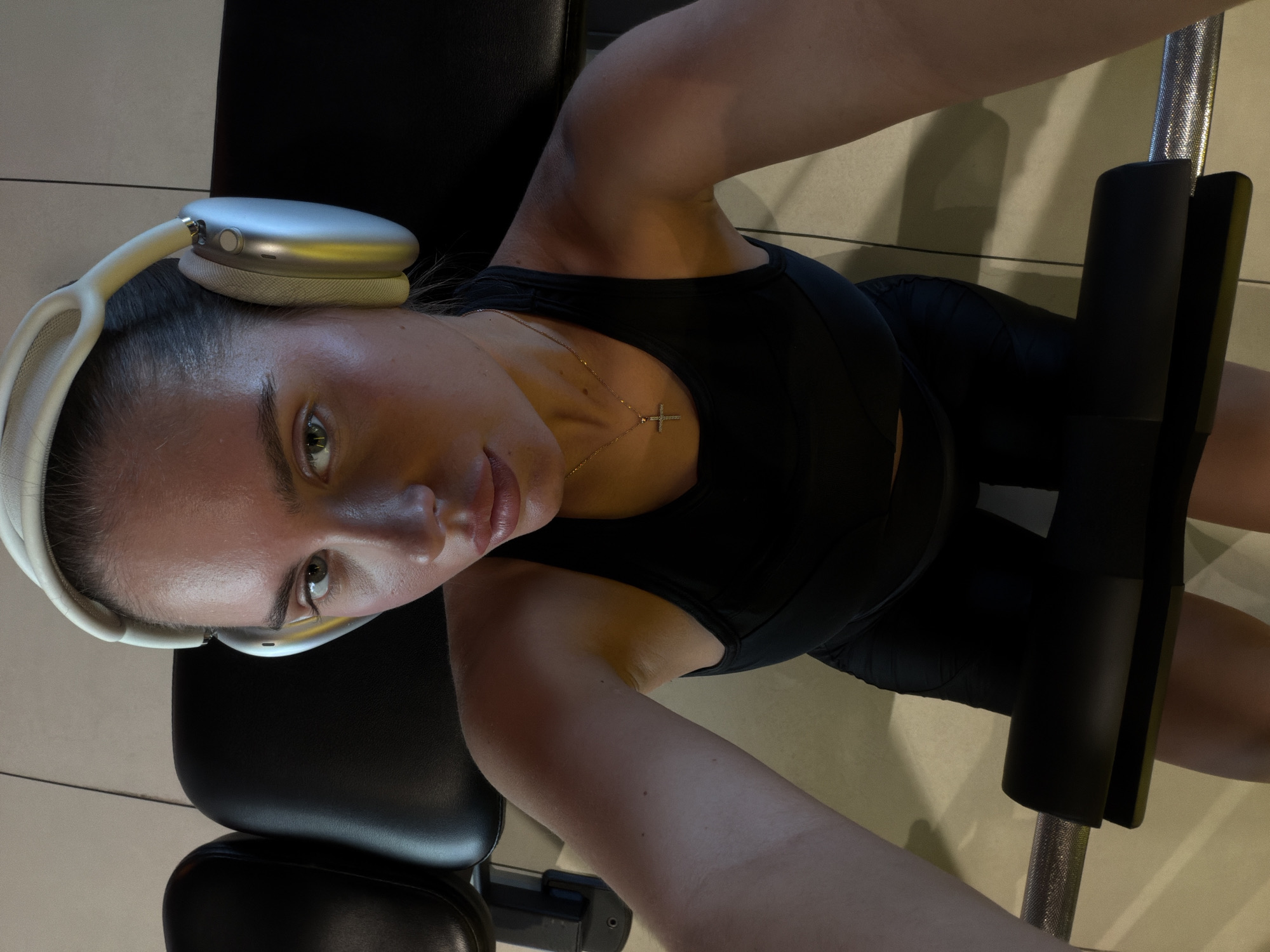 A woman wearing white headphones and a black athletic outfit is lying on a black leather bench while performing an exercise on a leg press machine. The woman is wearing a necklace with a cross pendant. The setting appears to be a fitness center with tiled floors. The location in Krasnoyarsk, Russia cannot be determined from this image.