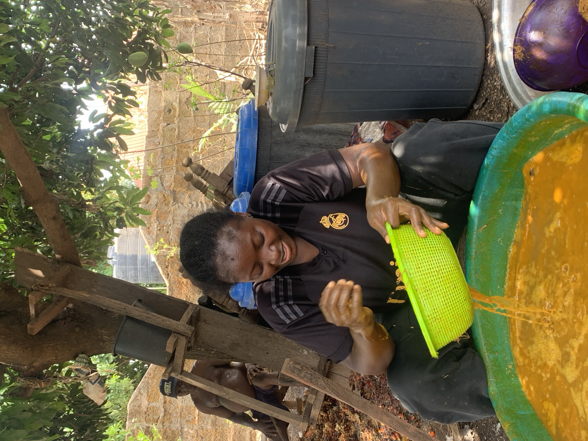 A woman is shown in Akwanga, Nigeria, processing something (likely palm fruit) using a green strainer over a large basin. She is wearing a black shirt with a logo and has dark hair. She is seated among other containers and possibly working with another person seen in the background. The setting appears to be outdoors, possibly in a yard, given the presence of a tree and other natural elements.