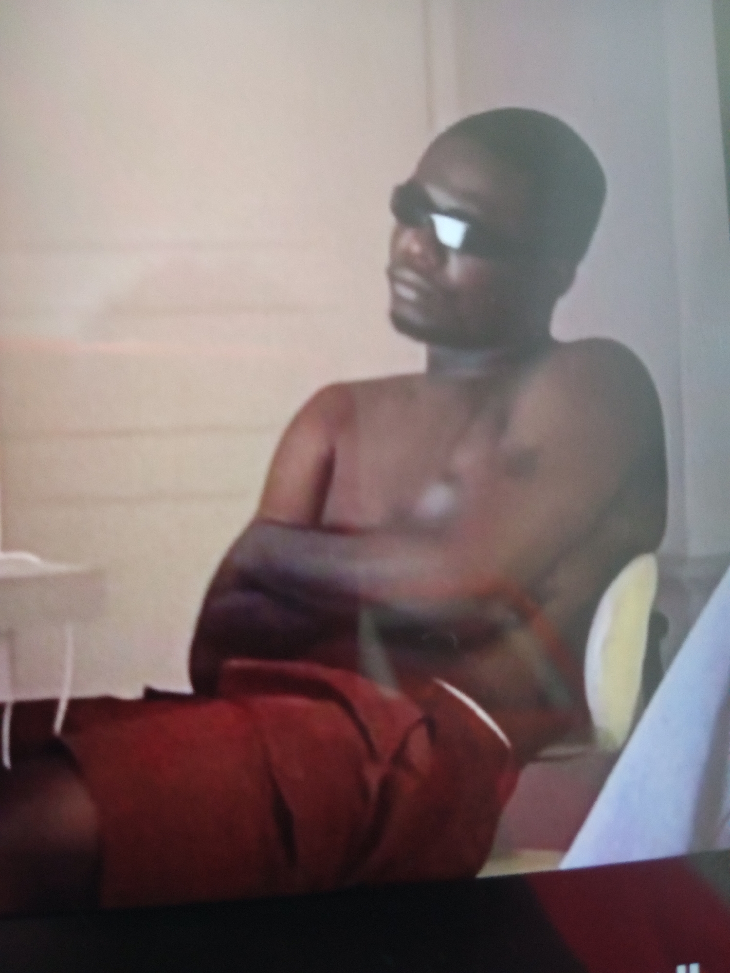 A man wearing sunglasses and red shorts is sitting with his arms crossed. The environment appears to be indoors, with a light-colored wall and some furniture visible. The image quality is low, making it difficult to discern finer details. The location is Jalingo, Nigeria.