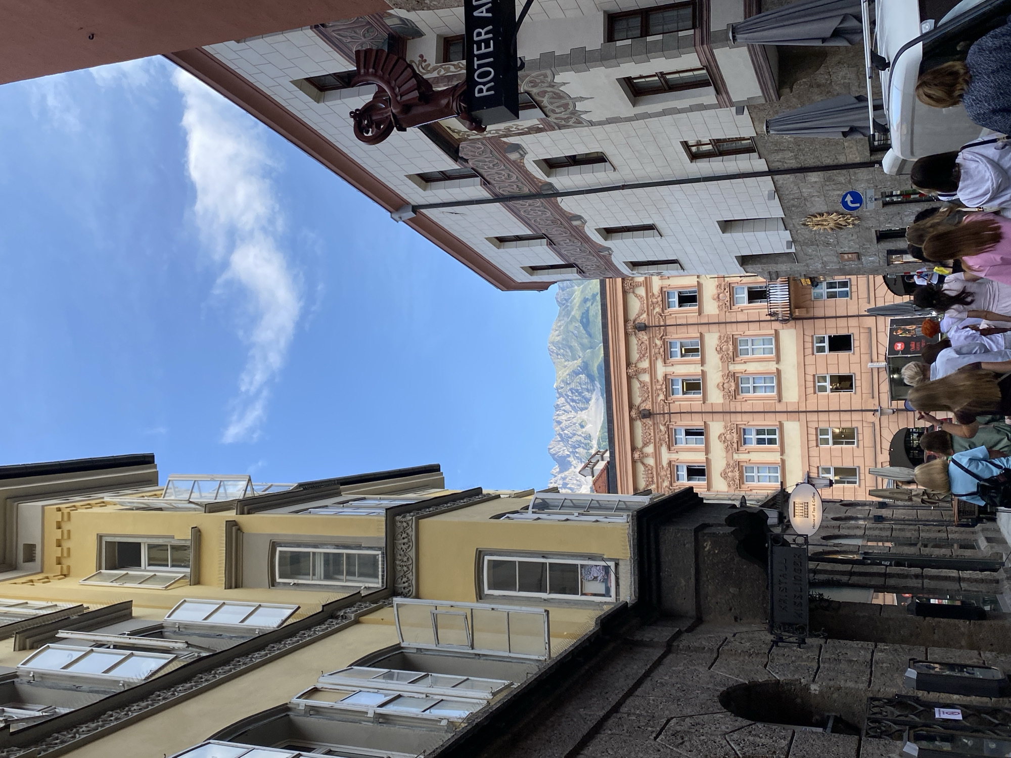 The image captures a street scene in Innsbruck, Austria, with a clear blue sky and a few scattered clouds. The architecture is a mix of old-world charm and more modern elements. On the left, a tall yellow building with multiple open windows and traditional shutters dominates the foreground. Its facade features intricate decorative elements along the roofline. Adjacent to it is a darker, stone-built structure with arched doorways.
On the right, a building with a white tiled facade and reddish-brown trim stands prominently. A decorative sign with a red eagle-like sculpture and the text "ROTER AD" is affixed to its upper part. Further down the street, another building painted in soft pink hues with white ornate details and numerous windows is visible. In the background, the majestic, snow-capped peaks of the Alps rise above the rooftops.
A group of people, mostly women, are walking away from the camera, appearing to be tourists exploring the city. They are casually dressed, suggesting a pleasant day for sightseeing. The street is paved, and some closed umbrellas are leaning against a building on the right. The overall impression is a vibrant European city with a strong historical character and a stunning natural backdrop.