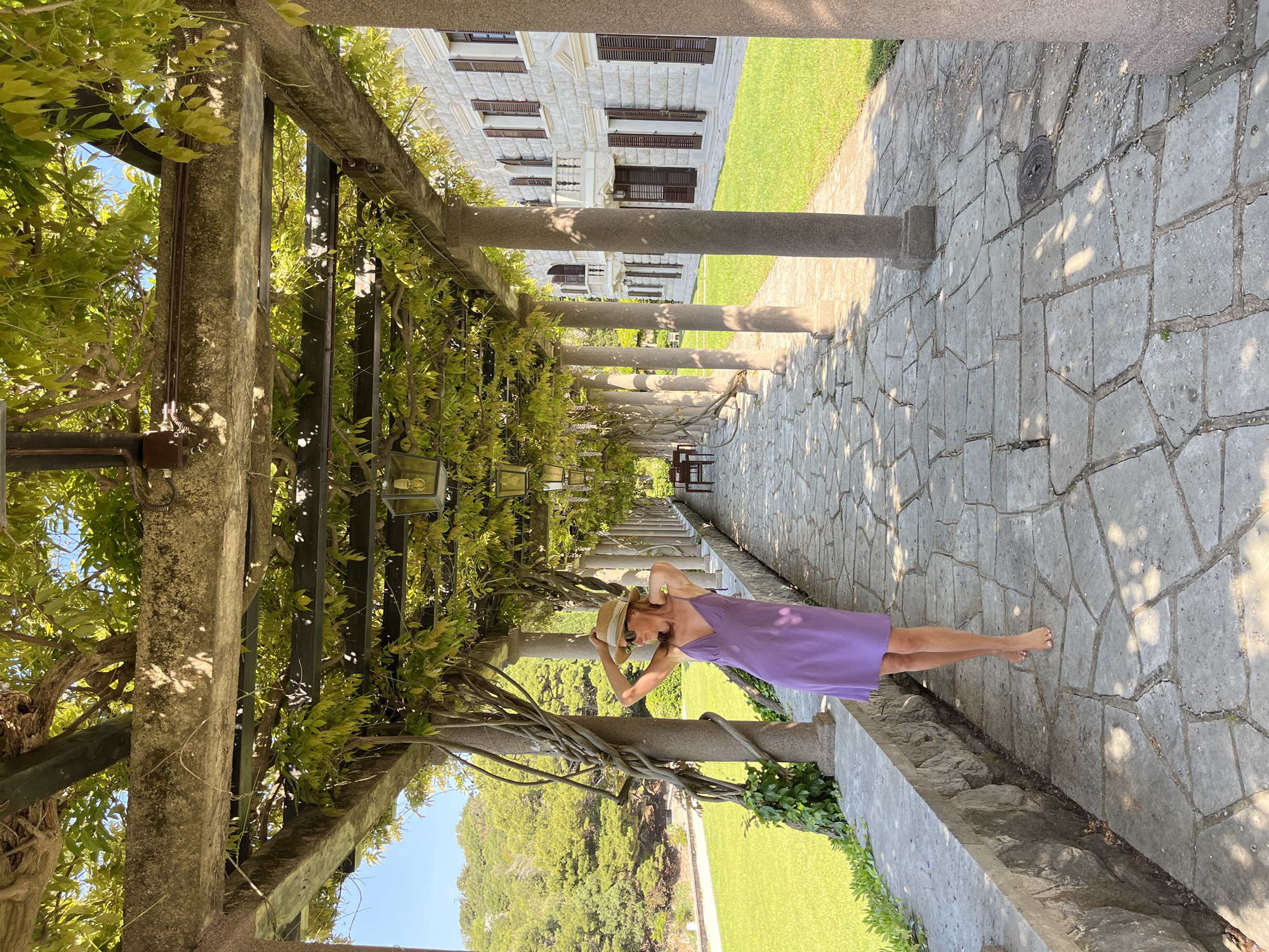 A woman wearing a purple dress and a straw hat stands in a pergola-covered pathway on a sunny day, likely in Sveti Stefan, Montenegro, based on the architecture and landscape. The pathway is constructed of irregularly shaped stones, and the pergola is supported by stone pillars adorned with climbing vines and leaves. A manicured lawn and trees are visible on either side of the pathway. In the background, a multi-story building with brown shutters and white stone facade is seen. The woman is standing barefoot with her hands on her head, as if adjusting or holding her hat.