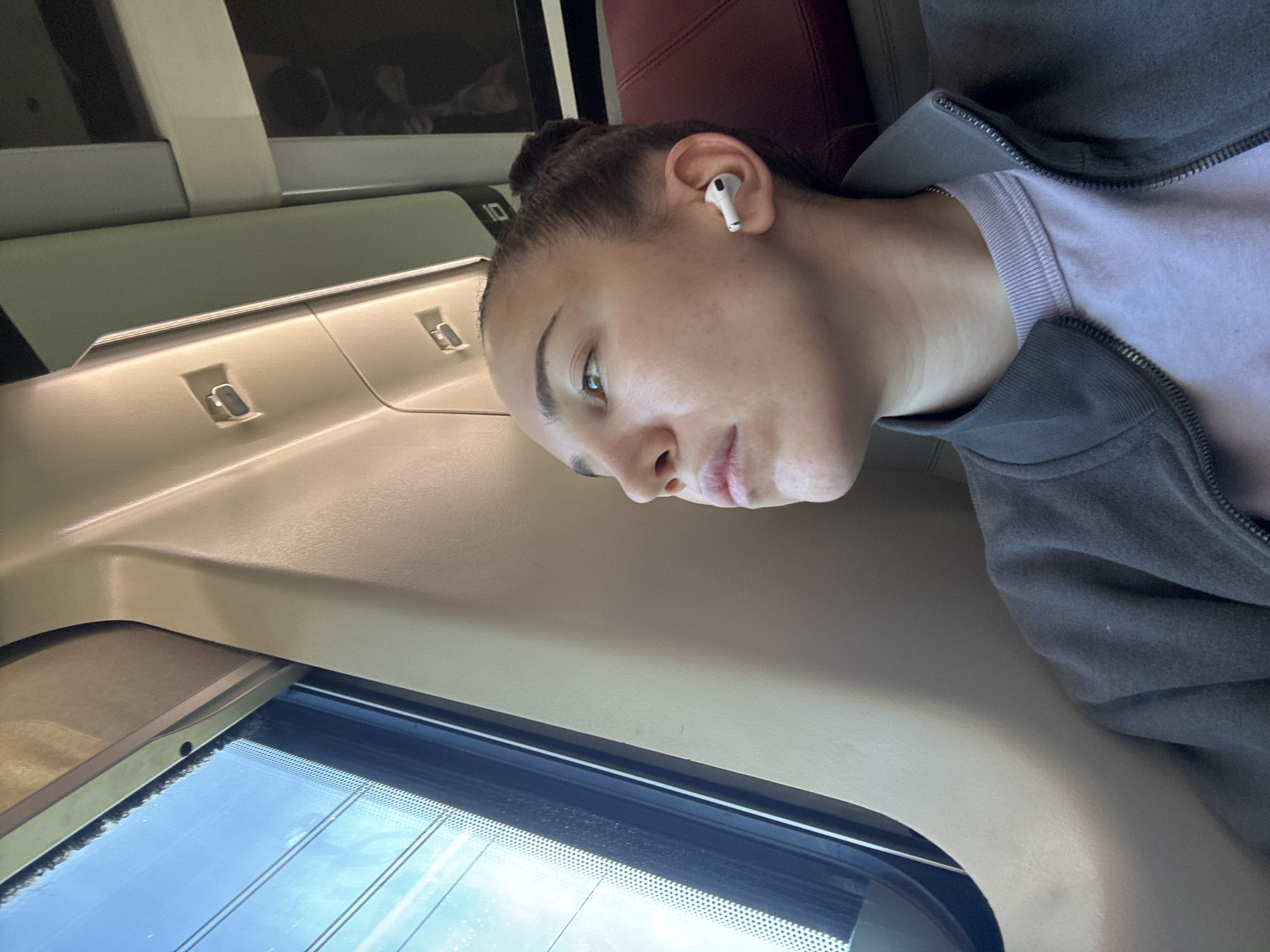 A woman with fair skin, wearing an AirPod in her left ear, is seated by a train window. She is wearing a light purple t-shirt and a grey zip-up sweatshirt. The scene appears to be inside a passenger train. The window shows a glimpse of the outside environment. The train's interior includes overhead compartments and red seating. There is no direct evidence indicating the location is Collevecchio, Italy.