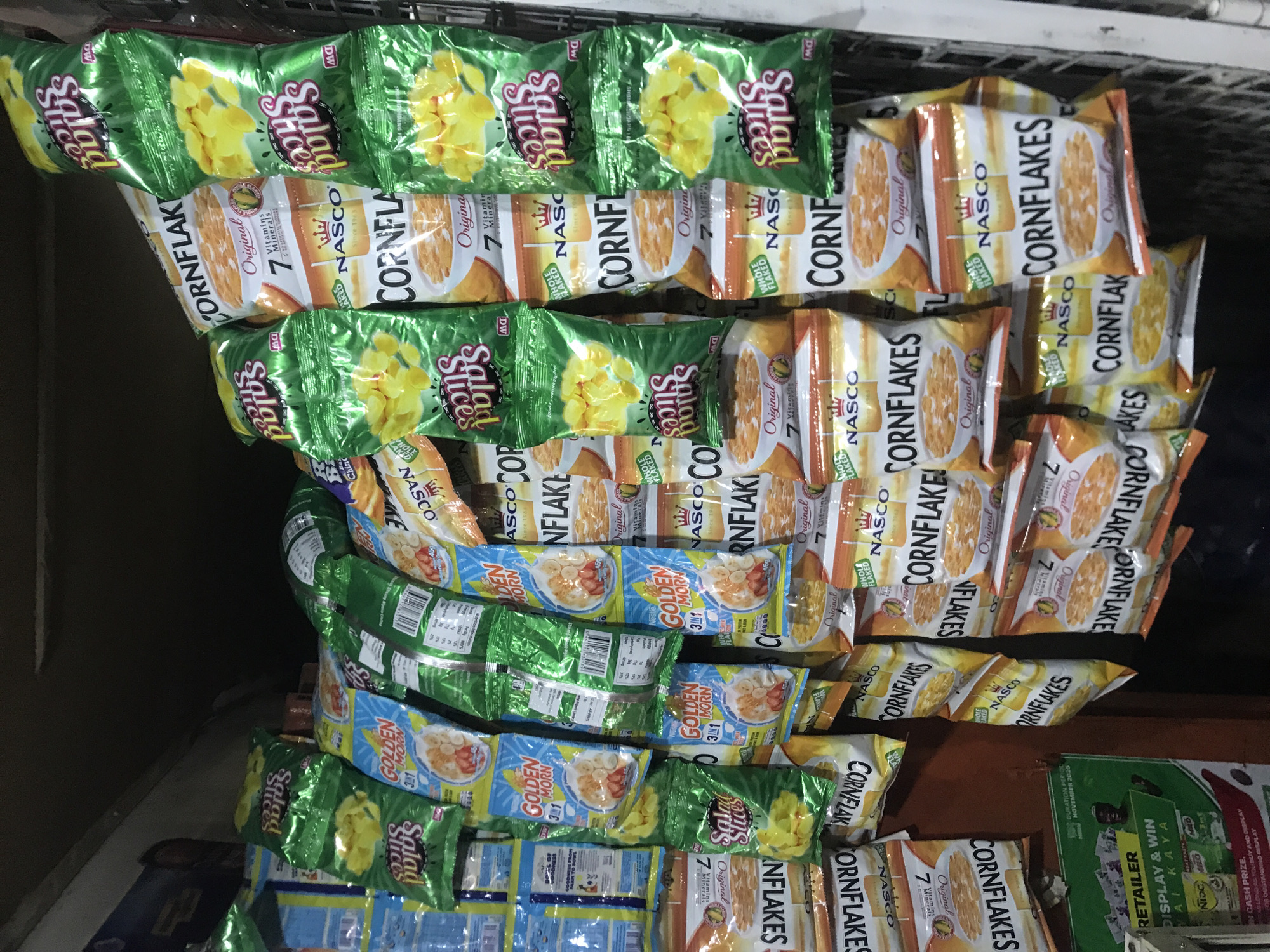 This is a close-up, eye-level shot of a retail display inside a small shop in Kabong, Nigeria. The display is packed tightly with various bagged food items, predominantly cereals and snacks. The items are hung from hooks or shelves, creating a dense visual texture.
Prominently featured are numerous bags of "NASCO CORNFLAKES" in orange and white packaging. These bags depict a bowl of cornflakes and highlight "Original" flavor and "7 Vitamins." Another recurring item is "Salad Slices" in green packaging, showing a pile of yellow, crispy snack pieces. There are also bags of "GOLDEN MORN" cereal, with a blue and yellow design, featuring a bowl of cereal with fruit. Smaller, less distinct blue and yellow packets are also visible.
In the lower left corner, a green promotional poster for "RETAILER DISPLAY & WIN" is partially visible. It includes text about "CASH PRIZE," "DISPLAY AND WIN," and "OUTSTANDING DISPLAY." A small image of a man and a Milo tin are also present on this poster.
The overall environment appears to be a dimly lit interior, likely a small grocery store or market stall. The background is dark and indistinct, suggesting a narrow space or a shadowed area of the shop. The lighting is focused on the products, making them the primary subjects. The arrangement of goods suggests a busy point of sale where a variety of packaged foods are offered.