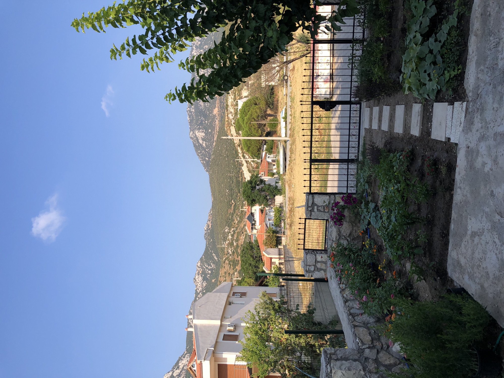The image depicts a residential area in Karaburun, Turkey, under a clear blue sky with a single wispy cloud. In the foreground, a garden with a variety of plants, including flowers and what appear to be vegetable plants like pumpkins, is visible. A stone wall borders the garden, and a black metal gate with decorative scrollwork stands at the entrance to a pathway made of stepping stones. Beyond the garden, a paved area with parked boats is visible. In the midground, several houses with red-tiled roofs are situated among green trees. A larger white building with brown window shutters is prominent on the left. The background is dominated by rugged, mountainous terrain with sparse vegetation. The overall scene conveys a peaceful, rural atmosphere in a coastal town.
