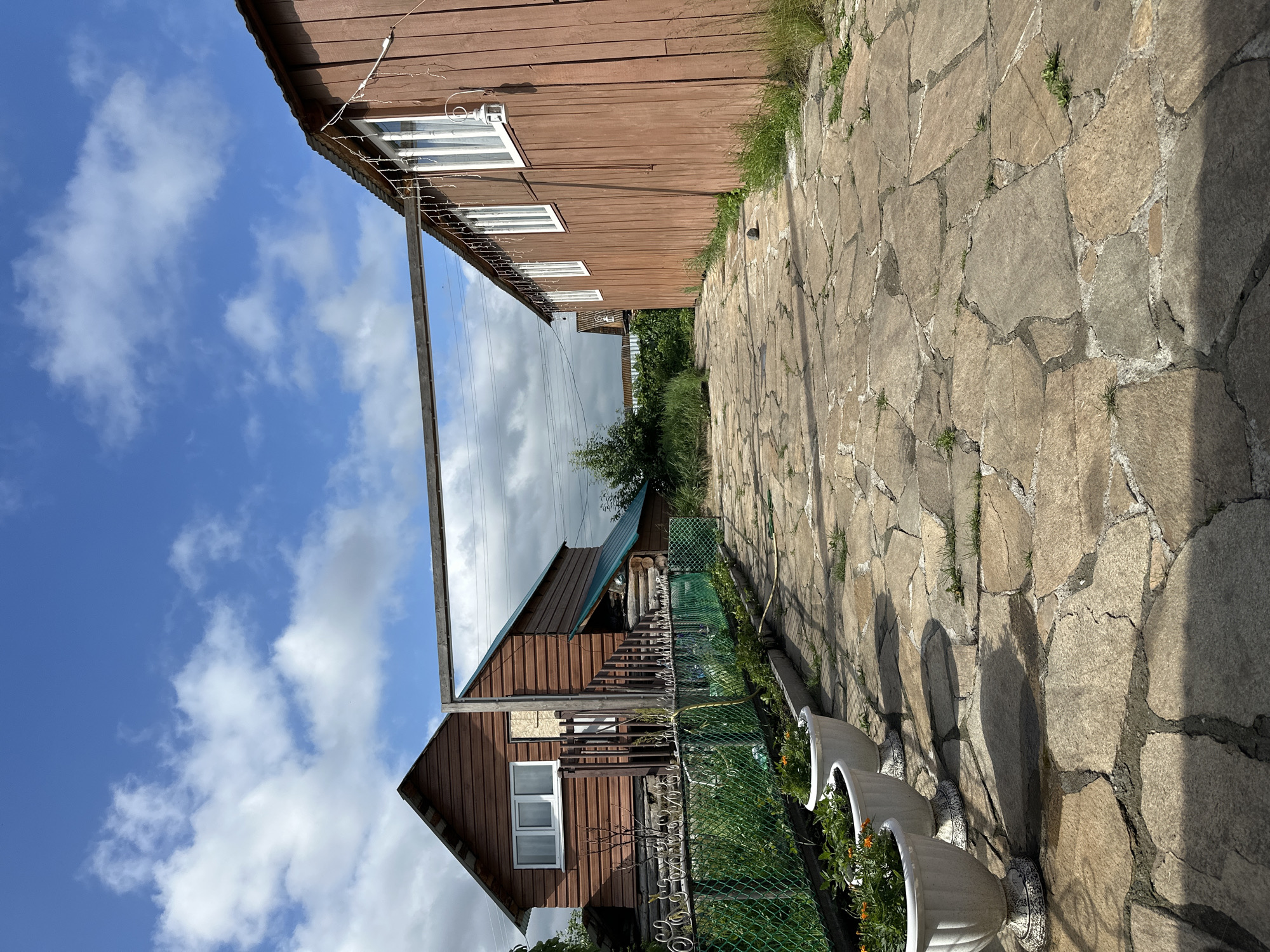 The image shows a stone pathway leading between two wooden buildings in a rural setting under a bright blue sky with scattered clouds. On the left, a two-story wooden house with a gabled roof and white-framed windows is visible, with a green fence and planters in front. On the right, a long, single-story wooden building with white-framed windows runs along the pathway. Decorative string lights are attached to the eaves of the building on the right. The pathway itself is made of irregularly shaped flagstones. Lush green vegetation and trees are present in the background, suggesting a garden or natural area. The overall impression is that of a rustic, possibly residential, property.