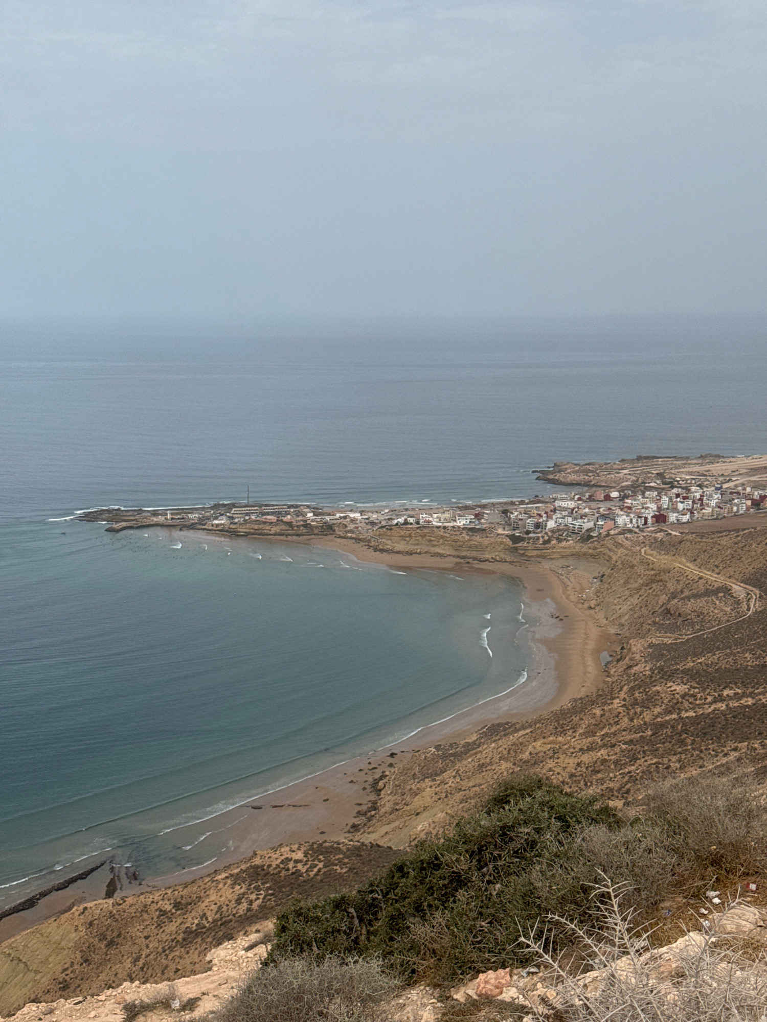 An aerial view captures the coastal town of Imsouane, Morocco, under an overcast sky. The town, characterized by its white buildings with red roofs, is nestled along a rocky coastline that curves into a sandy bay. The ocean, a deep blue, meets the shore with gentle waves. The surrounding landscape is arid and sparsely vegetated, with dry brush and rocks dominating the hillsides that slope down to the beach. A dirt road winds its way through the terrain, connecting the town to the higher ground. The water appears calm, with small whitecaps visible near the shore, and several surfers are scattered in the water, indicating a popular surf spot. The overall impression is one of a serene, remote coastal community in a rugged environment.