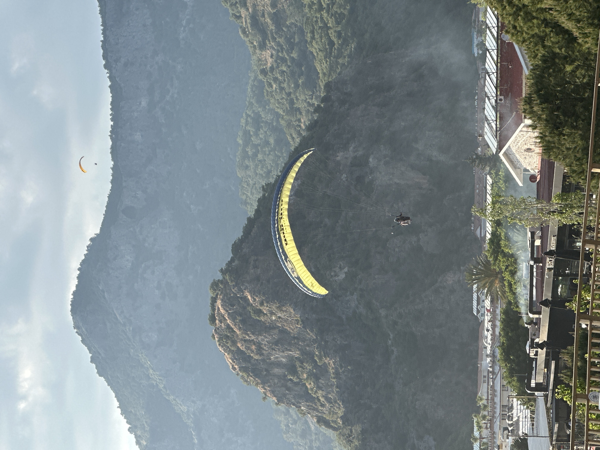 The image shows paragliders flying near a mountain in Fethiye, Turkey. One paraglider, with a yellow and blue canopy, is closer to the foreground, while another is visible in the distance. Lush greenery covers the mountain slopes. Buildings are visible along the bottom right.