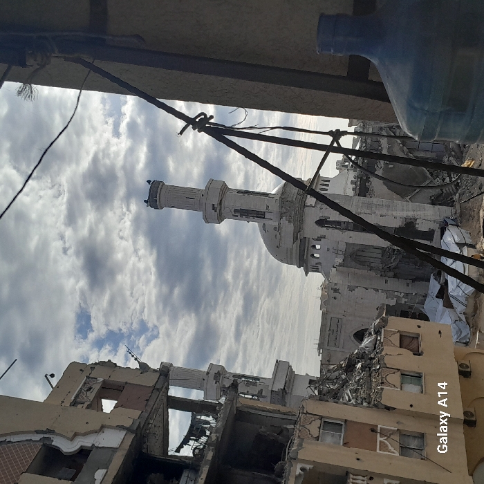 The image, taken in Khan Yunis, Palestinian Territory, depicts a scene of extensive urban destruction under an overcast or partly cloudy sky during daylight hours. The photograph appears to be taken from an elevated position, possibly from the roof or upper floor of a building.
In the immediate foreground, dark wires and what looks like a structural beam or part of a railing are visible, partially obscuring the view. To the upper right (when upright), a large blue plastic water bottle is also present.
The mid-ground and background reveal a landscape marked by severe damage. Numerous buildings, likely residential or commercial structures, are heavily destroyed, with walls blown out, interiors exposed, and large amounts of rubble and debris scattered. Some structures show multiple floors with damaged facades and missing sections.
Dominating the center of the scene is a large mosque, appearing relatively intact compared to its surroundings, though signs of wear or minor damage might be present. The mosque features a prominent white or light-colored dome and at least one slender minaret topped with decorative elements. Its architecture stands out against the damaged urban fabric.
No people or direct activity are visible, suggesting the aftermath of a significant event, likely conflict or bombardment, given the widespread structural damage. The overall atmosphere conveyed is one of desolation and destruction.
In the bottom right corner of the image, the text "Galaxy A14" is visible, indicating the device used to capture the photo.