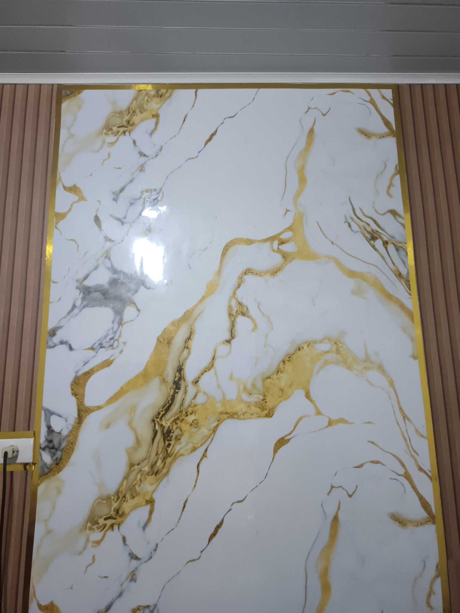 The image shows a large, decorative panel on a wall, featuring a white marble pattern with prominent gold veining. The panel is framed with a thin gold border. To the left and right of the central panel are vertical, wood-slatted wall sections, with the left section appearing a lighter shade of wood than the right. Above the decorative panel, a ceiling made of grey, ribbed panels is visible. In the lower left corner of the image, a white electrical outlet with a black plug and cord is attached to the left-hand wood-slatted section. The scene appears to be an interior, possibly in a residential or commercial space. The context provided by the prompt suggests this scene is located in Pangkalpinang, Indonesia, though no specific landmarks or contextual clues within the image confirm this location.