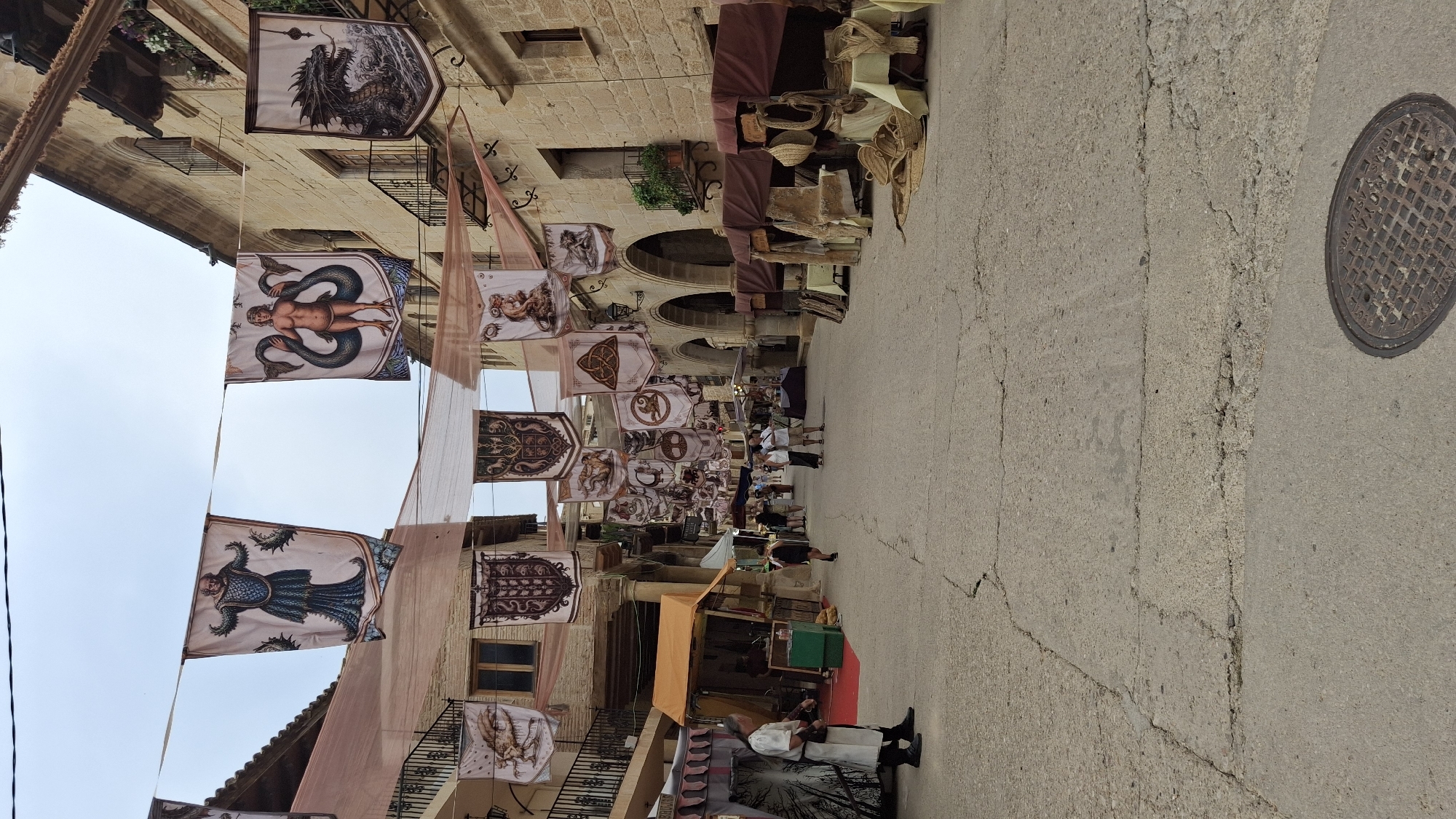 The image shows a street scene in La Fresneda, Spain, likely during a medieval fair or historical reenactment. Banners with various mythological creatures and emblems are strung across the street, suspended from the buildings. The buildings themselves are made of stone, with arched doorways and balconies. Along the sides of the street, market stalls are set up, displaying goods such as textiles and crafts. Several people are walking through the street, some dressed in historical costumes, including a man in a white tunic and dark trousers standing on the left side of the image. The ground is paved with cobblestones, and a manhole cover is visible in the lower right corner. The sky is overcast.