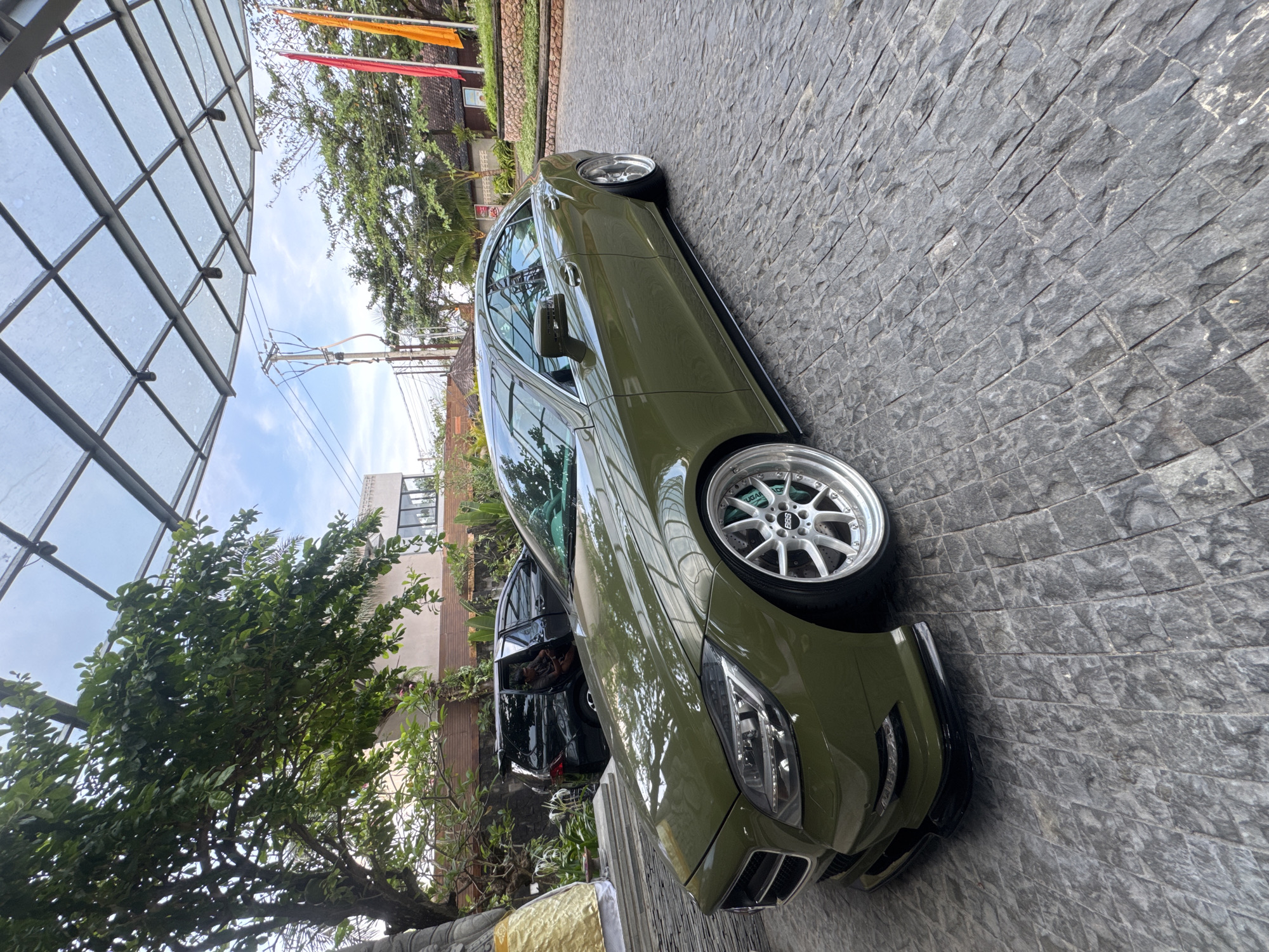 The image depicts an olive-green sedan parked on a cobblestone driveway. The car features custom wheels and appears to be modified. Adjacent to the sedan, partly obscured, is a black vehicle. Overhead, a modern glass awning structure is visible, reflecting the sky and surrounding vegetation. The scene suggests a location within a developed, possibly upscale area, with trees and buildings in the background. The image is taken in Kuta, Indonesia.