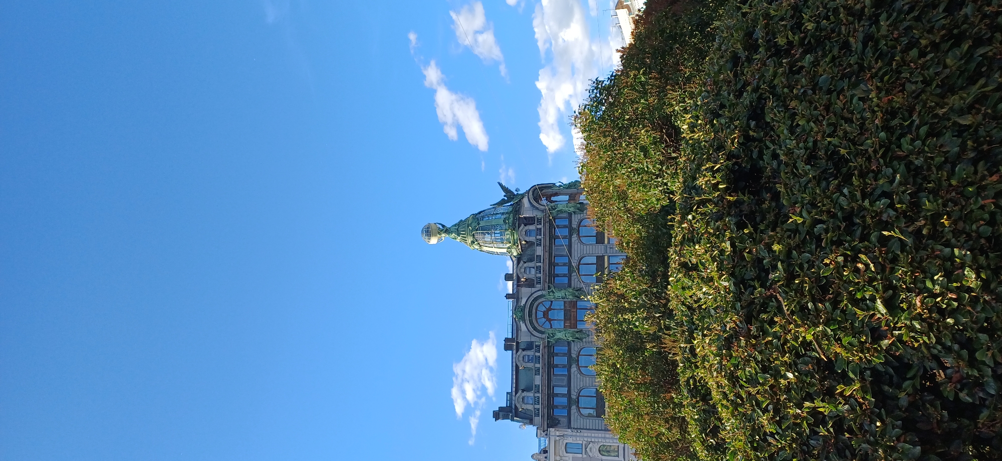 The image shows a view of a building with a distinctive dome, partially obscured by lush green foliage. The building features classical architectural elements, with arched windows and decorative sculptures adorning its facade. The dome is topped with a globe-like structure and features ornate metalwork. The sky above is a clear, bright blue, dotted with fluffy white clouds, suggesting a pleasant day. The sunlight indicates it is daytime, likely mid-morning or afternoon given the angle of the light. The overall scene evokes a sense of grandeur and historical significance, characteristic of European city architecture. The presence of dense, well-maintained greenery suggests a park or a well-landscaped area adjacent to the building. The image appears to be taken from street level, looking upwards towards the building and sky. There is no visible text in the image. The context provided places this scene in Saint Petersburg, Russia, a city renowned for its rich history and beautiful architecture.