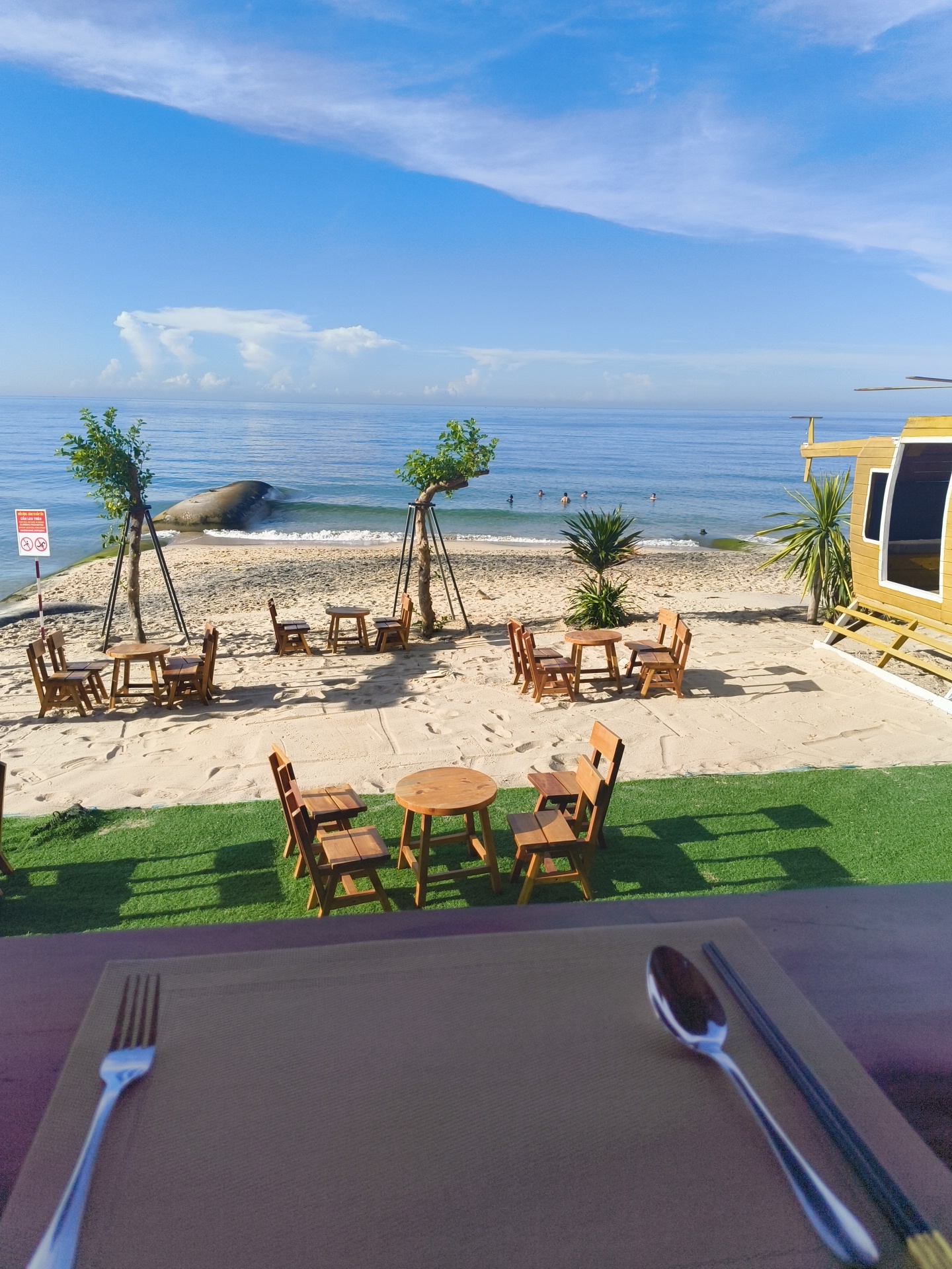 The image depicts a seaside dining area in Phu Thuy Ward, Vietnam, with the ocean in the background. In the foreground, a table is set with a placemat, fork, spoon, and chopsticks. The middle ground features a sandy beach with several sets of wooden tables and chairs, some shaded by small trees. Further back, people are visible in the shallow water, and a large rock formation is present near the shoreline. To the right, a yellow structure resembling a helicopter cockpit, possibly a unique seating area or part of a themed establishment, is visible. The sky is blue with scattered white clouds. The overall scene suggests a relaxed, outdoor dining experience by the sea.