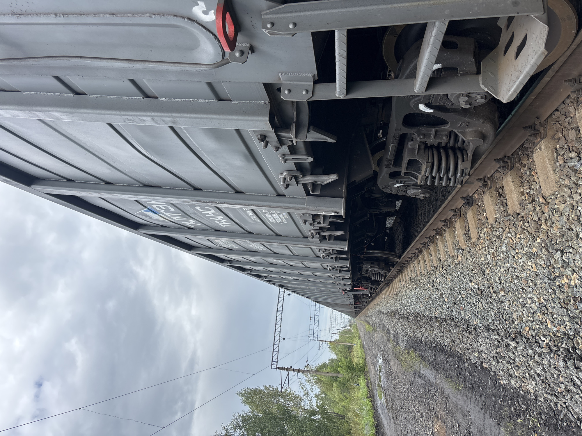 The image shows a close-up, low-angle perspective of a grey freight train on a railway track. The train cars are long and metallic, with visible corrugated sides. Some Cyrillic lettering is visible on the side of the car closest to the camera, including the word "ТРАНС". The train is situated on a gravel track, with some muddy ground and puddles visible to the left of the tracks, alongside a grassy embankment and trees. Overhead power lines and supporting structures for the railway are visible in the background, stretching into the distance. The sky is overcast with grey clouds. The scene appears to be a rail yard or a rural railway line.