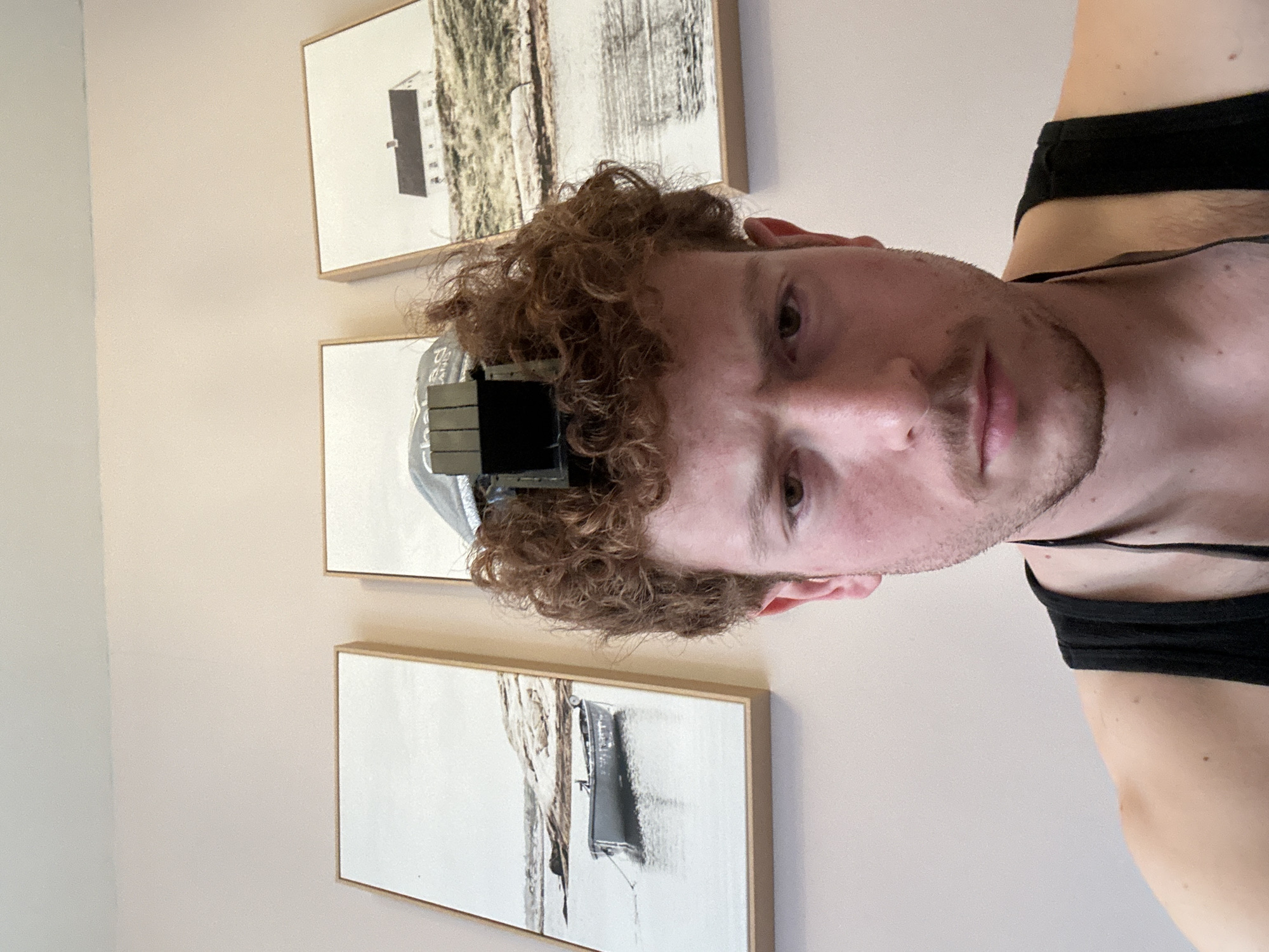 A vertically-oriented close-up shot features a Caucasian male with curly red hair, a light complexion, and a neatly trimmed beard. He is wearing a black tank top, and a gray hat with a black object attached to it is placed on his head. Behind the man are three framed pictures hanging vertically on a wall with a light pink hue. The artwork in the frames appears to be black and white landscapes or architectural images. The location of the scene is not discernible based on the provided image. The city and country are not identifiable from this image.