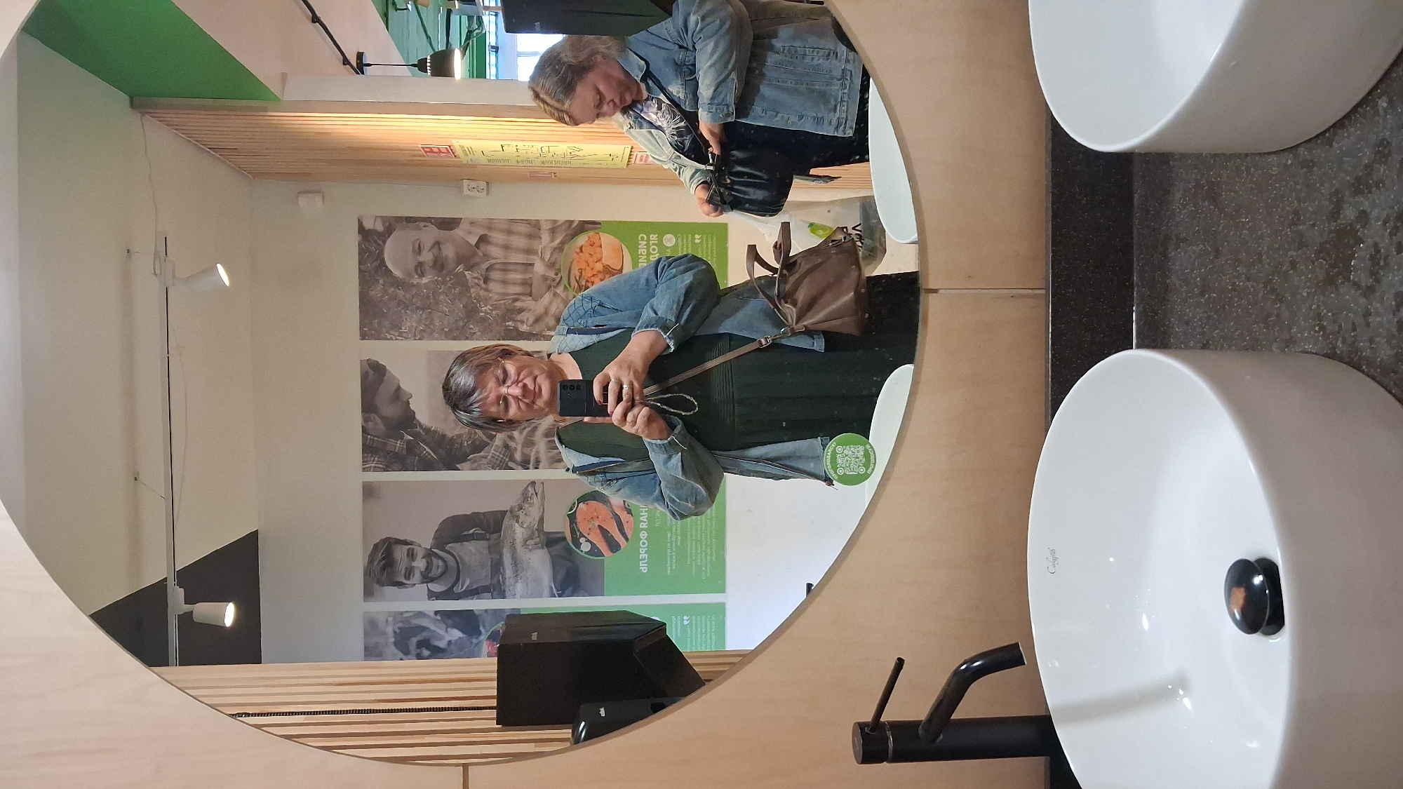 A woman is taking a selfie in a restroom mirror. She is wearing a dark green dress and a denim jacket. She has glasses and short, gray hair. A brown purse is slung over her shoulder. Behind her, another woman is looking into her purse. The restroom appears to be public, with two sinks and large posters on the wall behind the mirrors. The posters feature black and white photographs of people and food. The scene is well-lit by overhead lights.