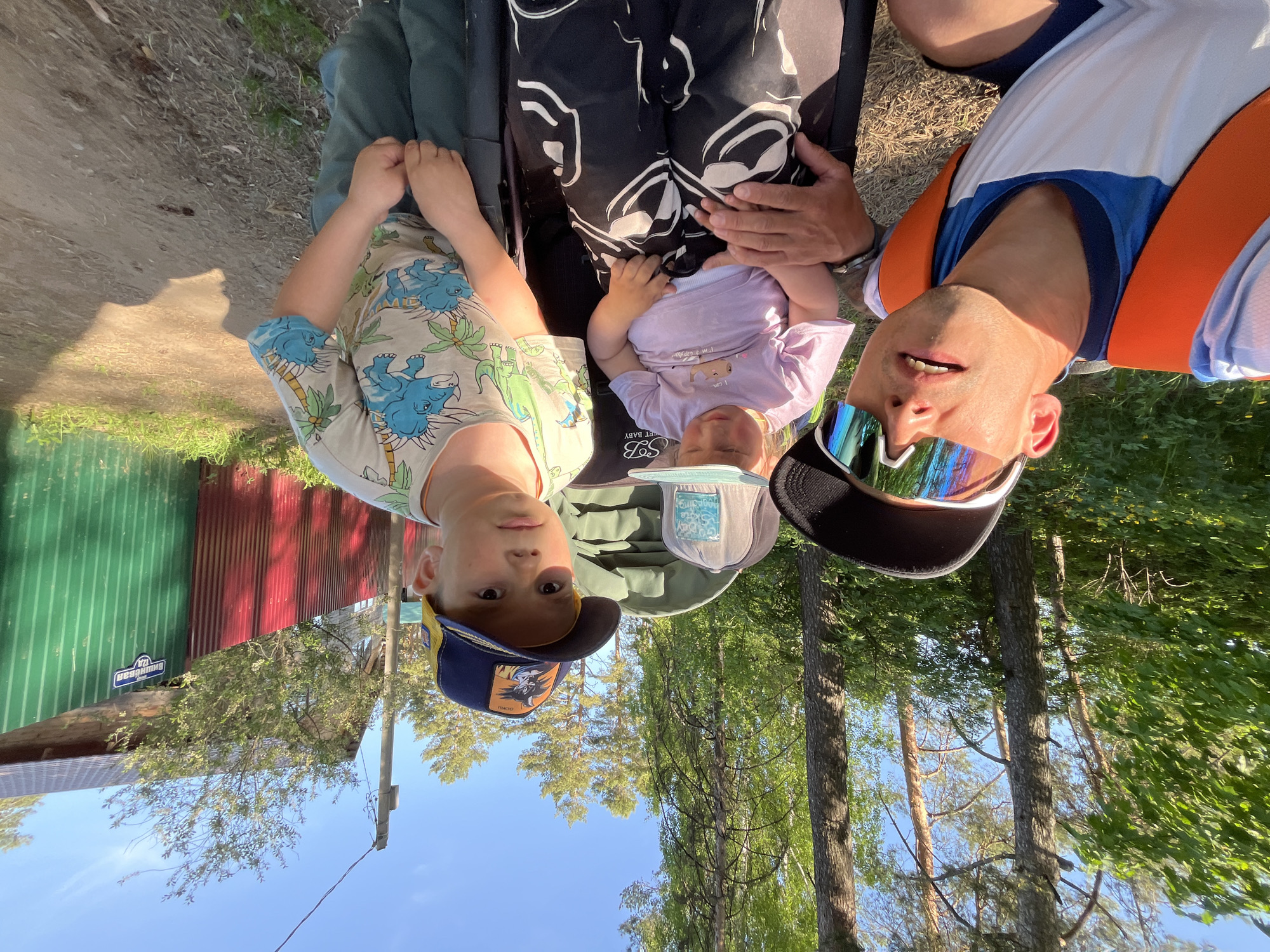 A man and two young children are positioned for a photo outdoors, with the camera facing them upside down. The man is on the right, wearing a white, blue, and orange top with sunglasses. The older child is to the left, wearing a patterned shirt and a baseball cap, and the younger child is in the center, wearing a light purple shirt and a bucket hat, lying in a stroller. The surrounding environment includes trees, grass, and part of a building made of corrugated sheet. The location is Chaikovsky, Russia.