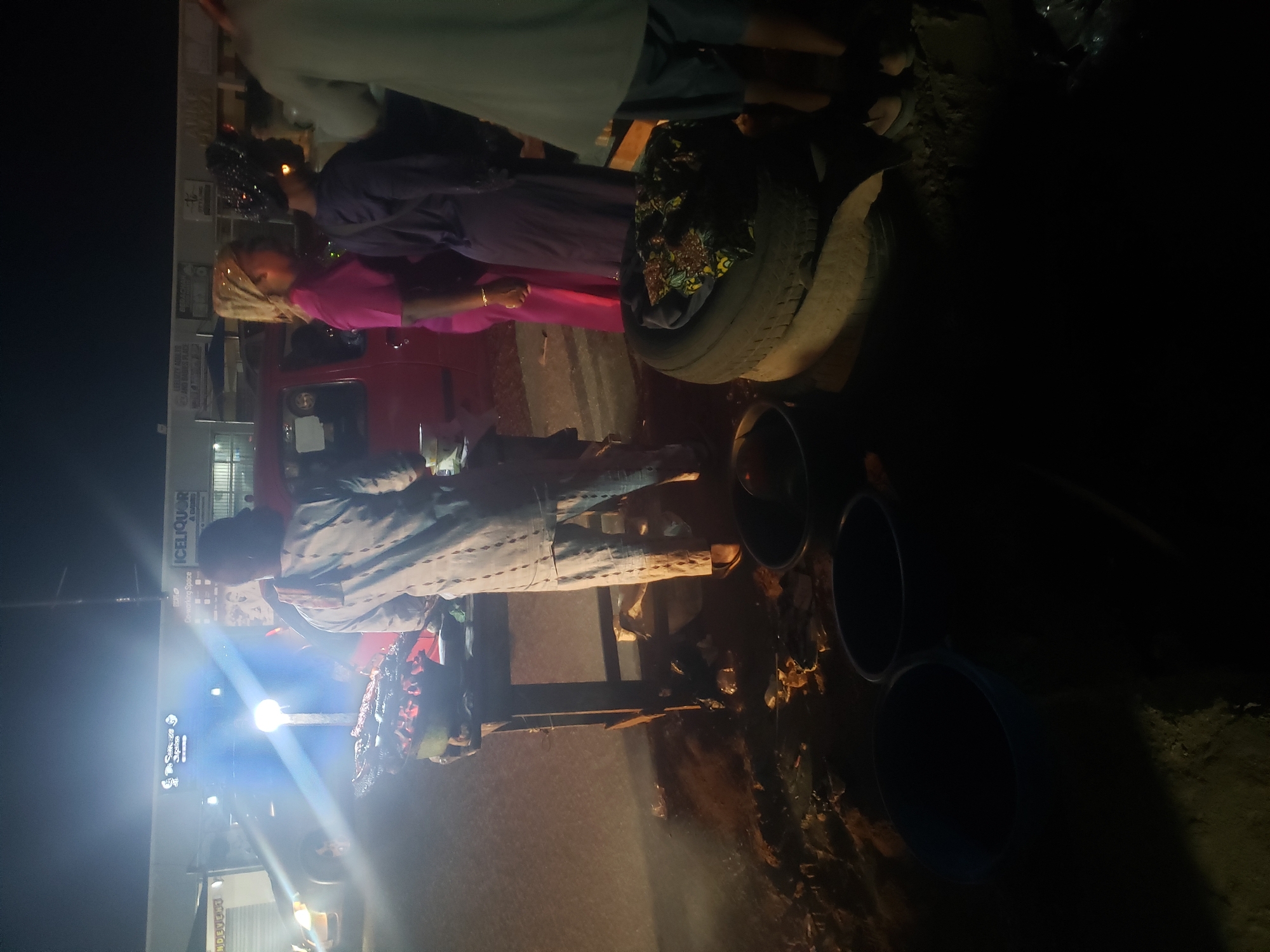 At night in Ibadan, Nigeria, a group of people are gathered around a street vendor. The vendor is positioned in front of a shop with signage for “ICELIQUOR” and “The Success Space”. A bright light shines from a vehicle near the vendor. A red vehicle is near the group. Some people are dressed in traditional African clothing, with one person wearing a pink outfit and gold headwrap. The vendor's setup includes black buckets, a wooden table, and what appears to be a tire being used as a surface. The ground is littered with debris.