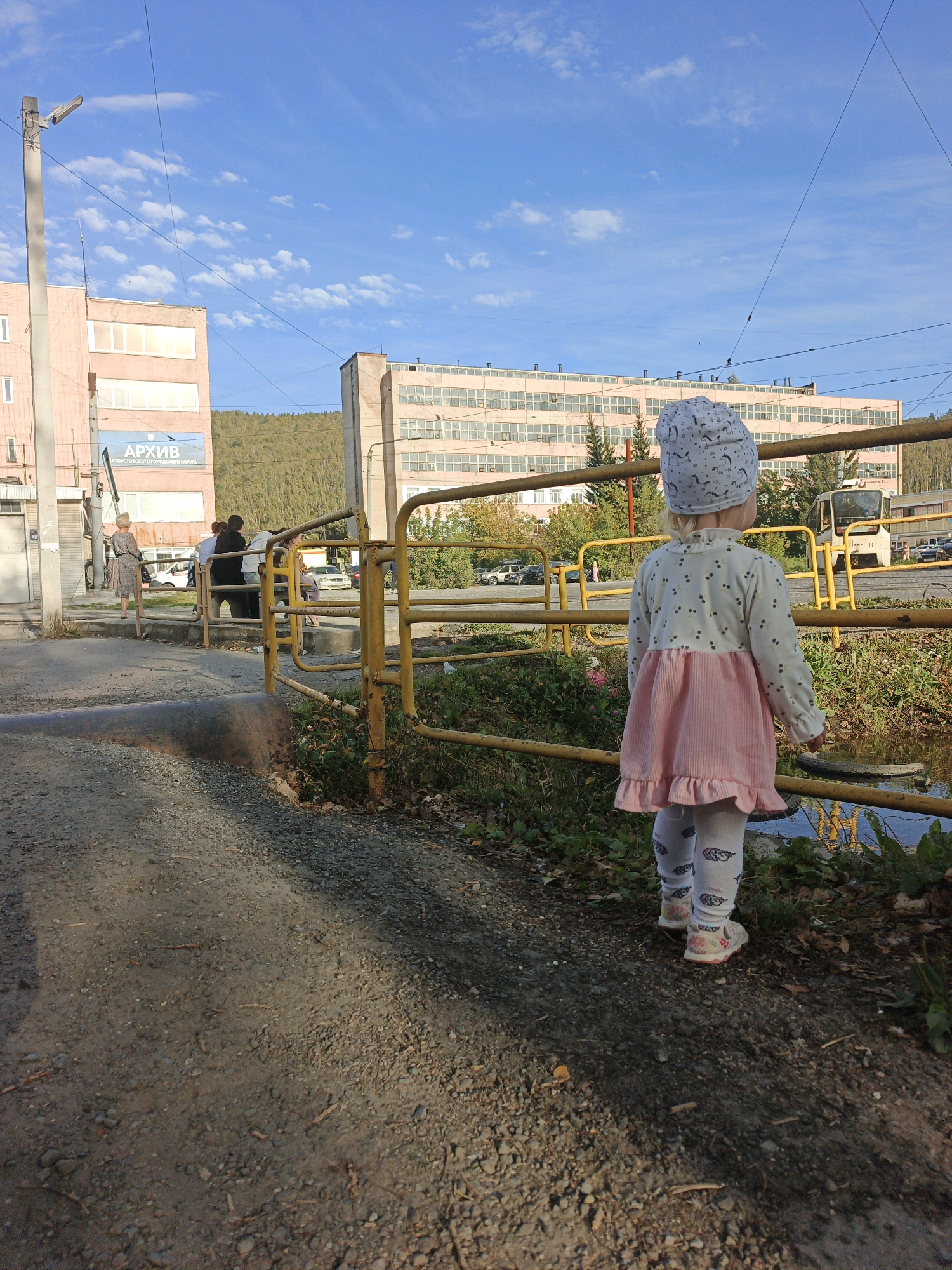 A young child, viewed from the back, stands on a gravel path facing away from the camera, toward a shallow body of water and a yellow metal barrier. The child wears a light-colored beanie with a dark pattern, a long-sleeved shirt adorned with cherry motifs, a pink corduroy dress, and white leggings with a feather pattern. The child is also wearing light-colored shoes with colorful accents.
In the background, across the water and barrier, is an industrial area. A large, light-colored, multi-story building with many windows dominates the left side of the background, featuring a blue sign with white Cyrillic text that reads "АРХИВ" (Archive). To the right of this building is another similar, longer industrial building. A light-colored bus is visible parked near the right industrial building. A tree-covered hill rises behind the industrial structures.
The foreground consists of a gravel path with scattered twigs and small stones. The sky is a clear blue with scattered white clouds. The scene appears to be set in Zlatoust, Russia, given the presence of Cyrillic text and typical industrial architecture found in Russian cities. The lighting suggests it is daytime, likely late afternoon due to the long shadows.