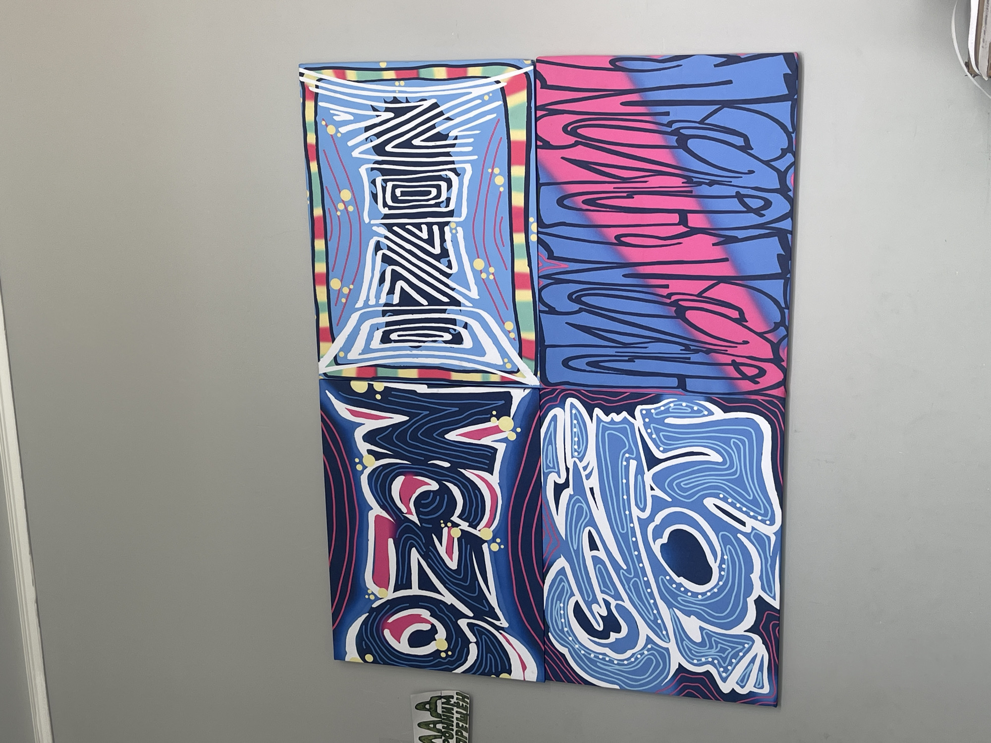 A four-panel graffiti-style artwork is displayed on a light gray wall. Each panel features the word "OZON" rendered in a different stylized font and color scheme. The top left panel has "OZON" in dark blue with pink and yellow accents. The top right panel shows "OZON" in white with a light blue background and colorful striped border. The bottom left panel displays "OZON" in various shades of blue with white outlines. The bottom right panel features "OZON" in dark blue outlines over a pink and blue gradient background. To the left of the artwork, a small white sticker with green lettering is affixed to the wall. The artwork is hung horizontally, forming a larger square composition.