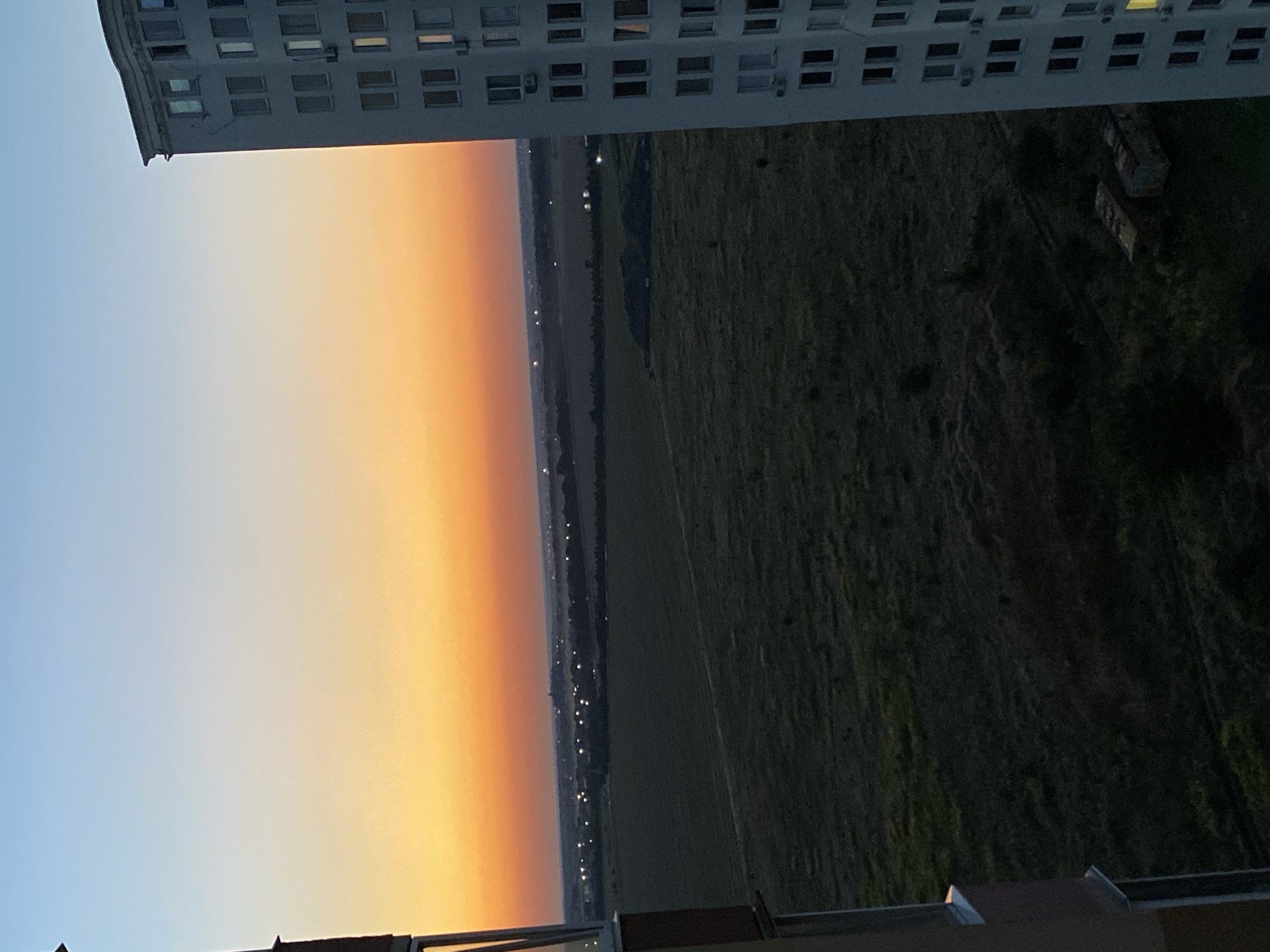 The image depicts a view from a high-rise building in Chornomorsk, Ukraine, during twilight. The sky transitions from a light blue at the top to a vibrant orange and then a softer yellow near the horizon. In the foreground on the right, a portion of a tall, modern apartment building with many windows is visible. On the left, a section of another building's facade and balcony railing is present. The middle ground shows a vast, dark green field or agricultural land, with some sparse vegetation. In the distance, a line of lights suggests a town or settlement. The overall atmosphere is serene and captures the quietude of early evening in an urban fringe area. There are no people visible in the image.