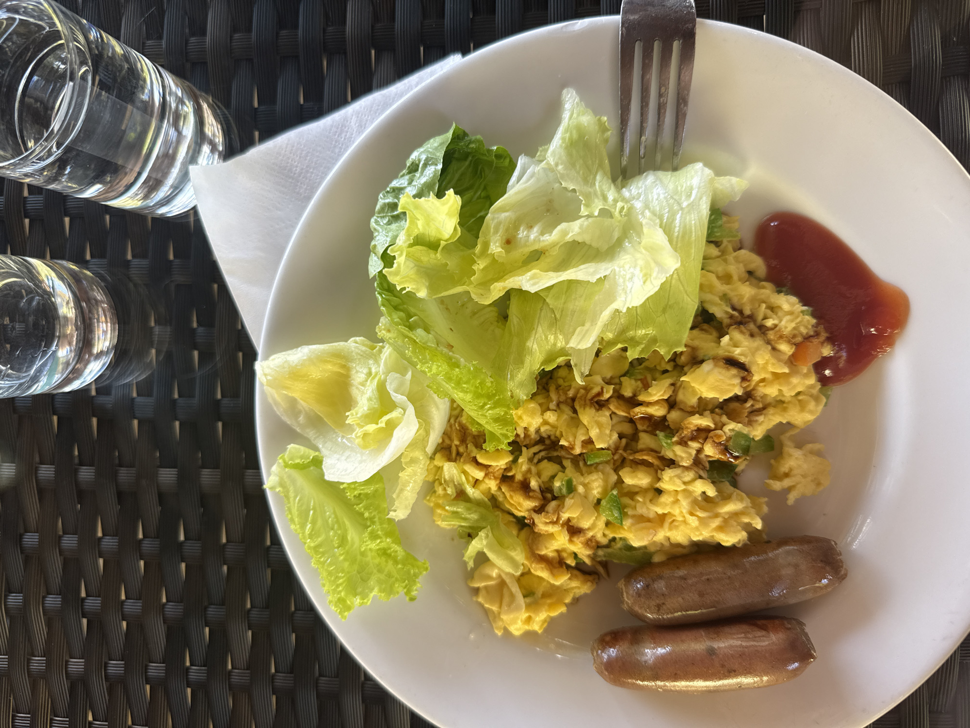 A plate of breakfast consisting of scrambled eggs, two sausages, and lettuce is presented on a table. The plate is white and sits on a dark, woven surface. To the right of the plate, there are two glasses of water. A white napkin is also visible. The setting appears to be outdoors, possibly on a patio or balcony, given the woven table and natural lighting. The background suggests an outdoor dining area, consistent with a resort or restaurant in Kuta Selatan, Indonesia. There are no people visible in the image.