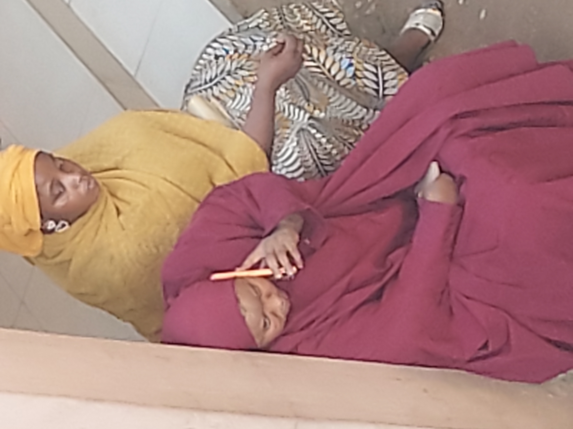 The image captures three individuals, likely women, resting or sleeping in an informal setting. The photo is rotated, showing the subjects lying horizontally across the frame.
On the left, a woman is visible wearing a bright yellow headscarf that covers her hair and neck, and a matching yellow garment. Her eyes are closed, suggesting she is asleep or deeply resting.
Between the woman in yellow and the one on the right, a third person is partially visible. This individual is covered by a patterned garment featuring white, grey, yellow, and black leaf-like designs. Their right arm is visible, resting on their body, and a foot wearing a simple light-colored sandal is seen towards the bottom right of the frame, indicating they are also lying down.
On the right side of the image, another woman is seen, covered entirely in a deep maroon or red head covering and body garment. Her face is visible, with eyes closed, also appearing to be resting or sleeping. Her right hand is placed on her chest, holding an orange rectangular object, possibly a mobile phone or a remote control.
All three individuals appear to be lying on a hard, light-colored surface, which could be a floor or a low platform. The background consists of simple, light-toned walls or structures, and the ground visible beneath them is a rough, light-colored surface. The overall atmosphere conveyed is one of quiet rest. There is no visible text in the image. The clothing styles suggest a cultural context consistent with the given location of Jos, Nigeria.