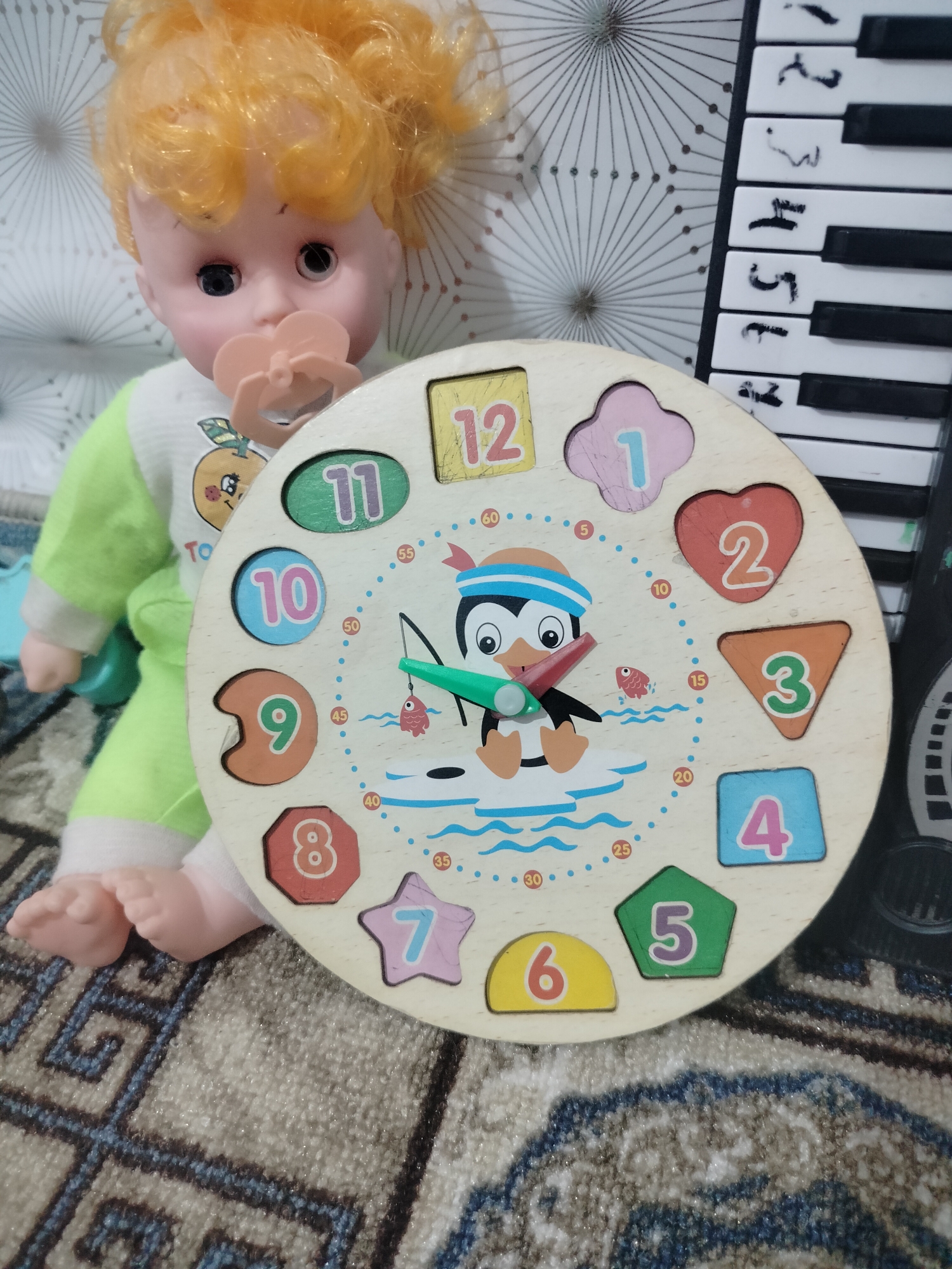 A doll with blonde hair and a pacifier is sitting on a patterned rug. In front of the doll is a wooden clock puzzle with numbers and shapes. To the right of the doll is a keyboard with black and white keys. The background consists of a white surface with a geometric pattern. The scene is indoors, likely in a domestic setting, but no specific city or country can be identified from the visual information provided. The doll is positioned with its legs extended, and the clock puzzle is tilted towards the viewer.