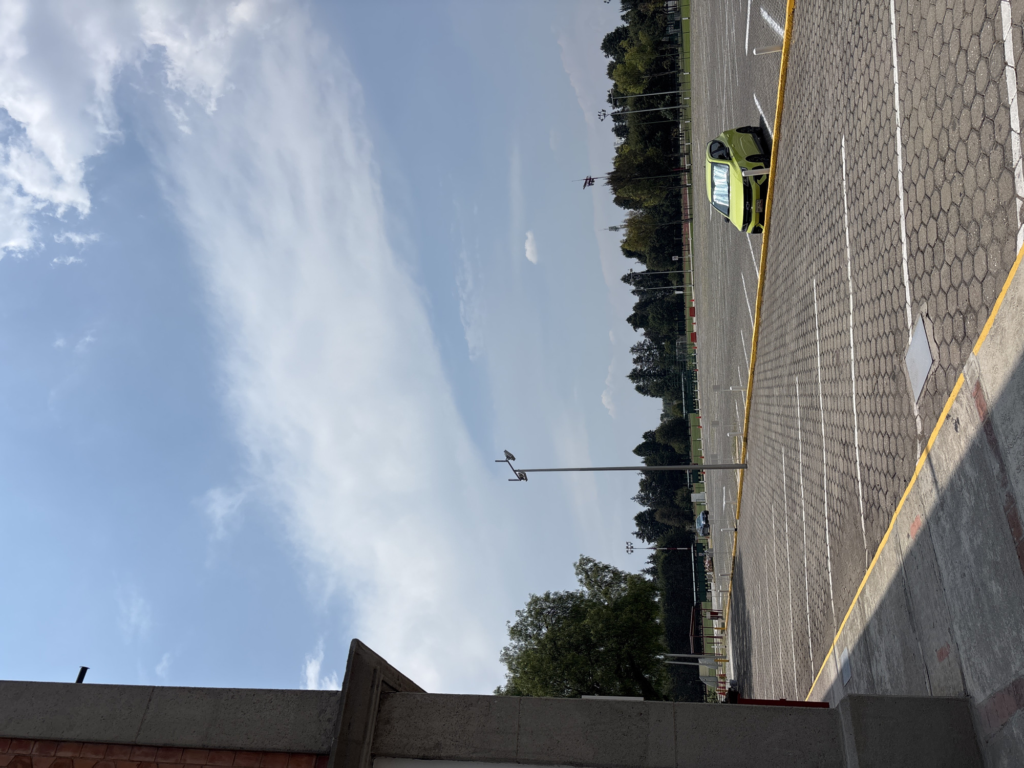 The image shows a partially filled parking lot in San Andrés Cholula, Mexico, on a clear day with scattered clouds. A lime green car is parked on the far right edge of the lot, near a low concrete barrier. The parking lot surface is gray with white parking lines and a yellow border. Beyond the parking lot are trees and some infrastructure, possibly a sports field or recreational area. The bottom of the image is obstructed by a low brick and concrete wall. Two light poles extend from the left side of the parking lot upward toward the sky.