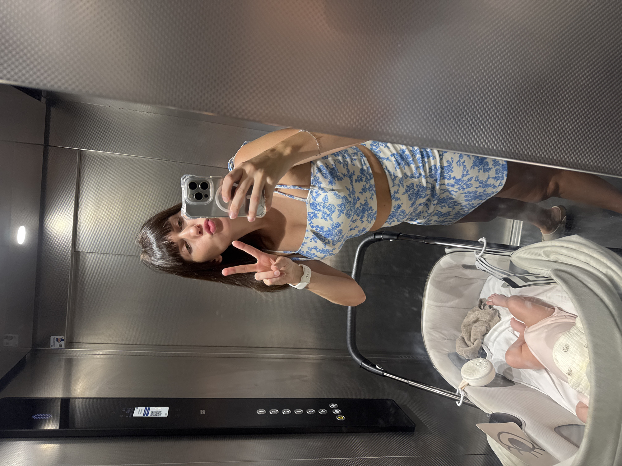 In an elevator with brushed metal walls, a young woman takes a mirror selfie. She is wearing a blue and white floral crop top and skirt, and her hair is dark and bangs-style. She is making a peace sign with her right hand and puckering her lips. To her left, a baby stroller is visible, containing a baby in a pink onesie. The elevator has a vertical control panel with illuminated buttons on the left side. The scene is likely in Isola delle Femmine, Italy, based on the context provided.