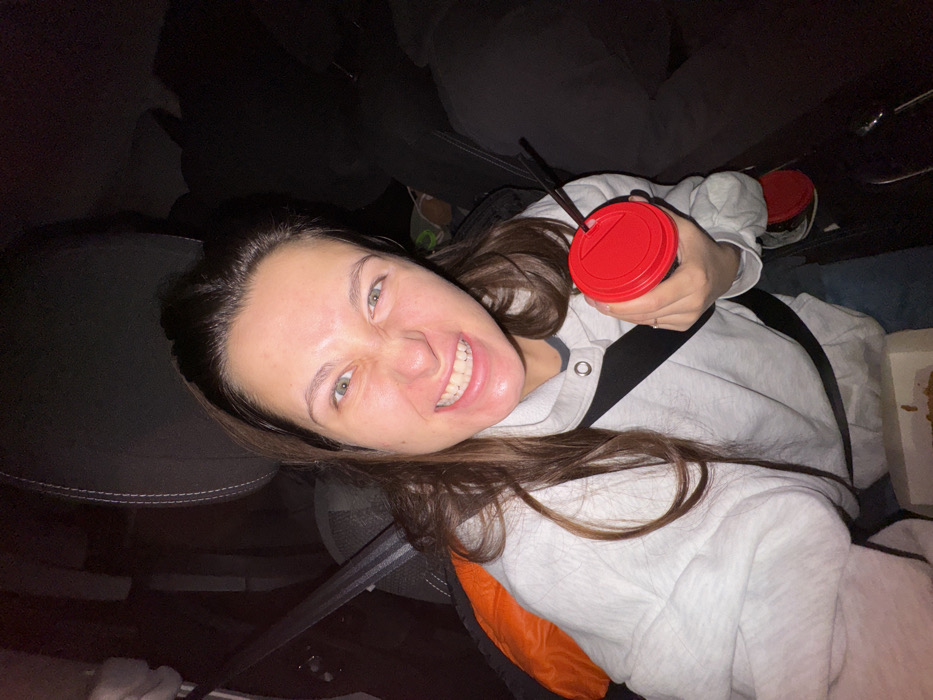 A woman grins at the camera, her teeth bared. She wears a light gray hooded sweatshirt and a seatbelt. She holds a red-lidded cup. A second red-lidded cup is in the background. She appears to be in a car, given the seatbelt and surrounding upholstery. The lighting is dim.