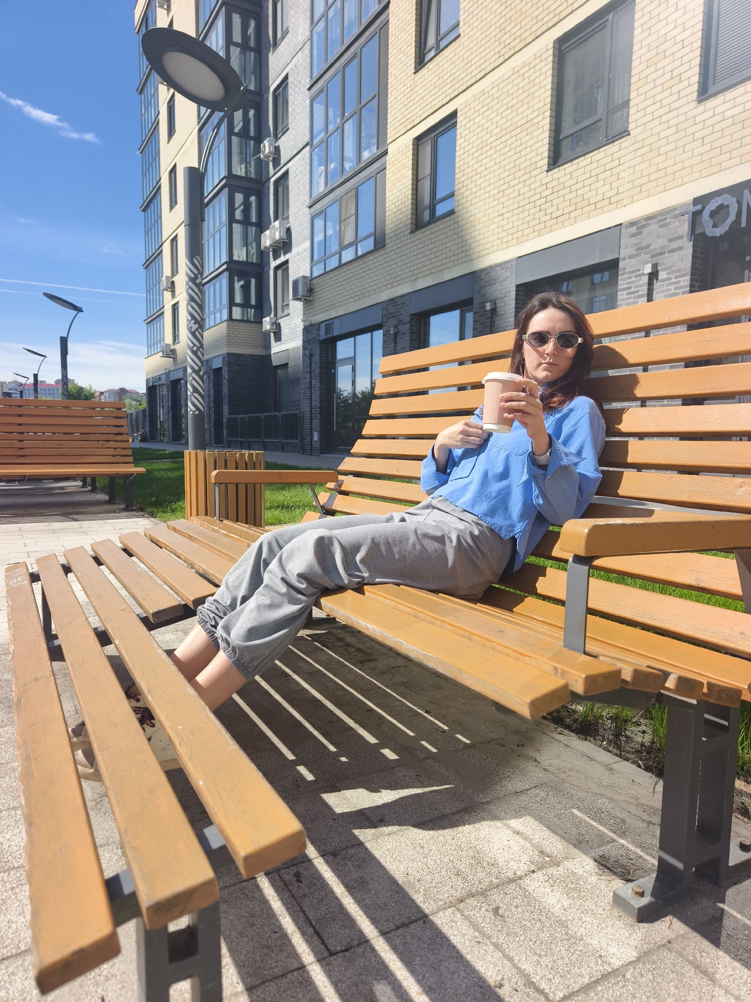 A young woman is captured in a full-body shot, relaxing on a slatted wooden bench in a sunny urban setting in Tyumen, Russia. She is seated with her legs extended along the bench, holding a light brown disposable coffee cup with a white lid in both hands. She is dressed in a light blue, long-sleeved button-up shirt, grey elastic-cuffed sweatpants, and stylish oval-shaped sunglasses. Her expression, partially obscured by the sunglasses, appears neutral as she looks towards the camera.
The scene is brightly illuminated by direct sunlight, casting strong, defined shadows from the bench and the woman onto the paved ground, indicating a clear, sunny day, likely mid-day or early afternoon. The sky above is a vibrant clear blue with a few wispy clouds. In the background, a modern multi-story residential building stands prominently, featuring large windows and a facade composed of contrasting light yellow brick and darker grey brick or paneling. Several external air conditioning units are visible on the building's exterior. Modern streetlights with large, flat circular luminaires are positioned along the paved area. Another identical wooden bench is visible further back on the left, alongside a patch of green grass. A partial word, "ТОН", and a circular logo are discernible on the darker facade of the building in the background.