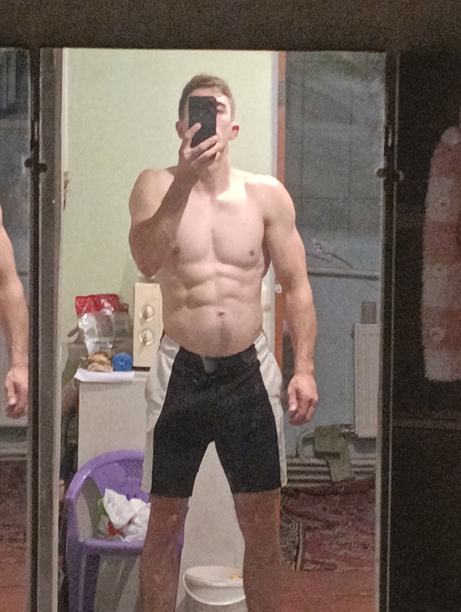 A young Caucasian male, appearing to be in his early twenties, stands shirtless in front of a mirror, taking a selfie with his smartphone. He has short, light brown hair and a lean, muscular physique, showcasing well-defined abdominal muscles and biceps. He is wearing black and white athletic shorts. The background appears to be an indoor setting, possibly a bedroom or bathroom, with a beige wall, a dresser with items on it, and a purple plastic chair. A portion of a closet or another mirror is visible to the left, and a dark, textured surface is on the right. The lighting is somewhat dim, suggesting it is either evening or the room is not well-lit. The image is taken from a slightly low angle, looking up at the subject. The location is identified as Fastiv, Ukraine.