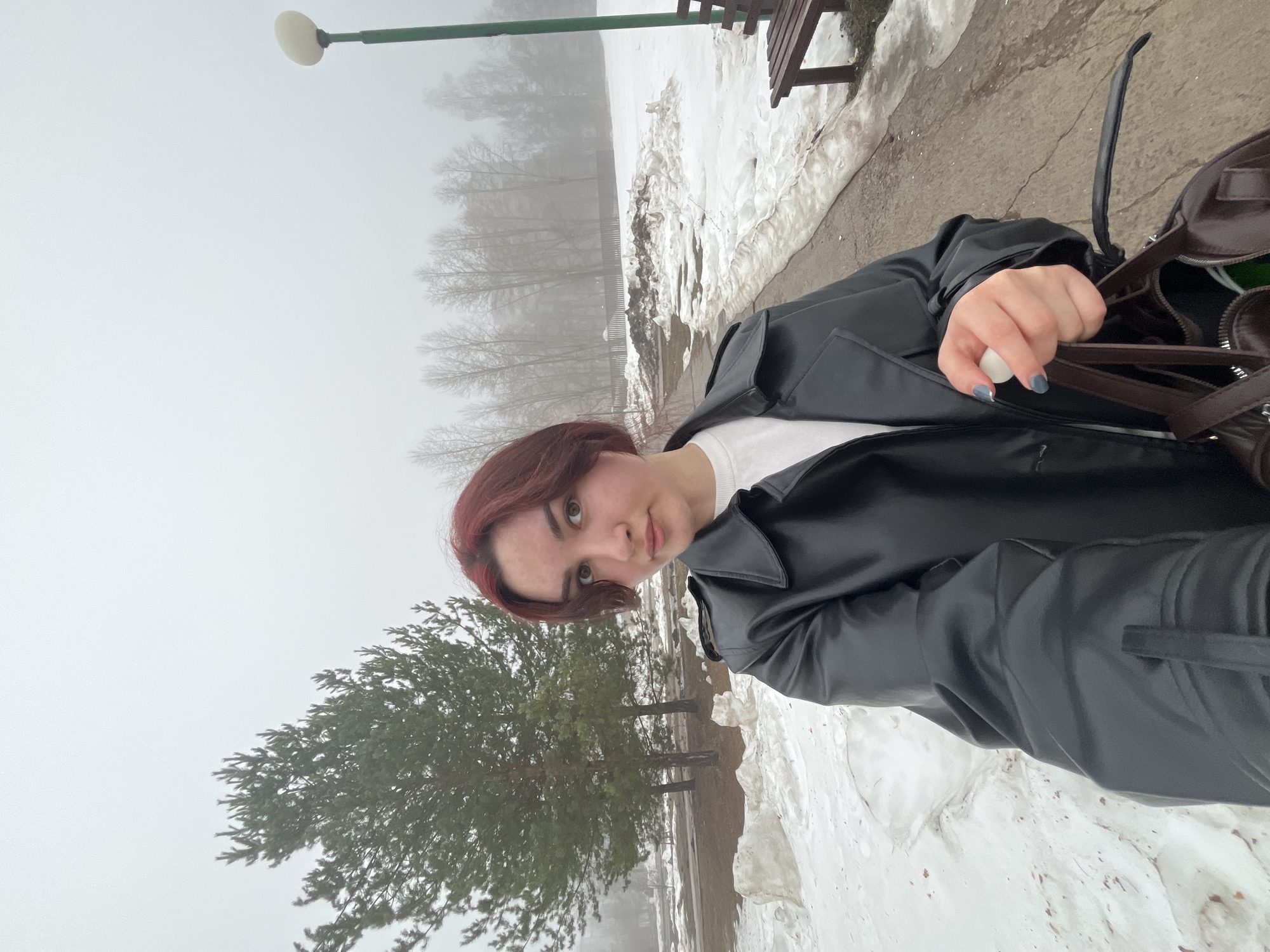The image depicts a woman in a black jacket and white shirt holding a brown bag outdoors. She has short, reddish-brown hair. The background shows a foggy or misty day in what appears to be a park setting. There are trees, snow patches, a lamppost, and a bench visible. Given the user's specification, it is likely the location is Neftekamsk, Russia. The woman appears to be taking a selfie or a photo, as the perspective suggests she is holding the camera herself.