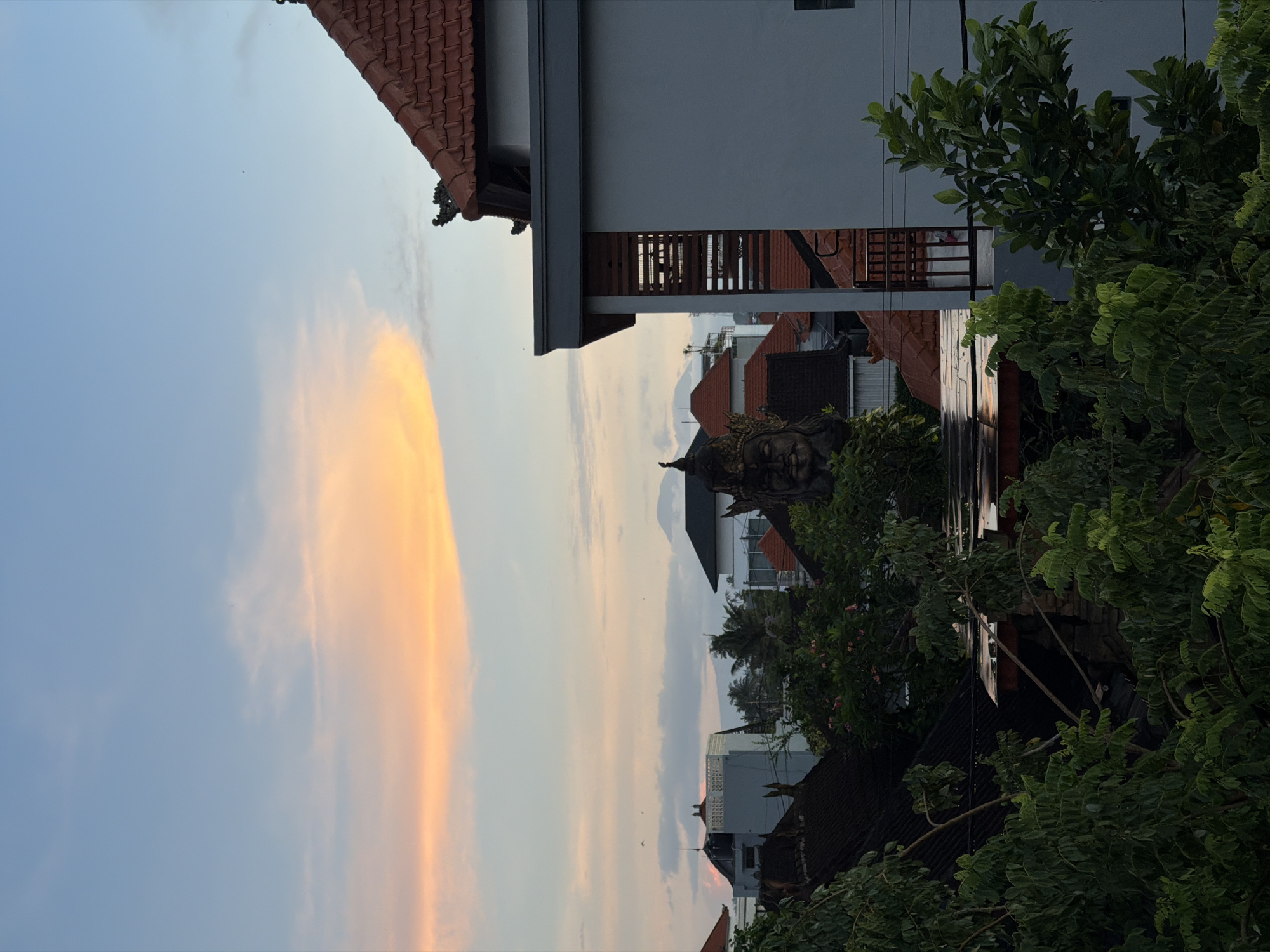 The image captures a scene in Mengwi, Indonesia, featuring a mix of residential architecture and natural elements. The sky shows a gradient of colors, with an orange-yellow light on the left side blending into a light blue. The top of a building with a red-tiled roof is visible in the upper left corner. Adjacent is a modern, multi-story structure with a light-colored exterior and slatted balcony railings.
In the midground, roofs of other buildings are visible, interspersed with trees and foliage. A statue with intricate detailing is partially visible amidst the foliage. The foreground features a dense cluster of trees with green leaves. The composition is oriented vertically, providing a view from an elevated perspective.