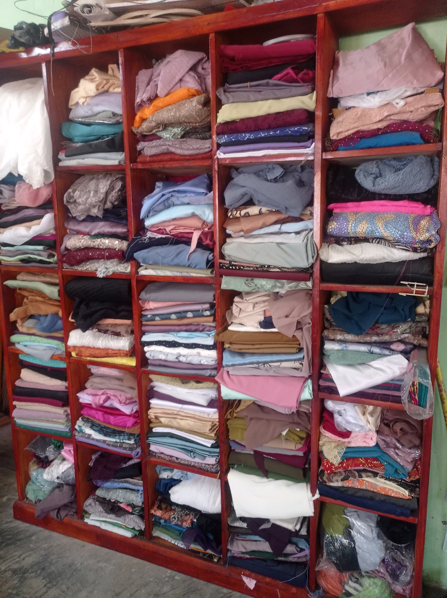 The image shows a wooden shelf unit filled with neatly folded fabric and clothing. The shelves are organized into multiple compartments, each containing stacks of various colors and textures of material. The unit appears to be in a room in Campang, Indonesia, based on the context provided. The lighting suggests it is daytime, and the environment seems to be a storage area or a workshop. There are no people visible in the image.