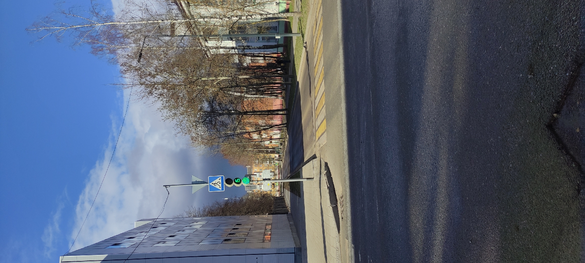 The image shows a street scene in Fryazino, Russia. On the left, there is a tall building with a reflective facade. A pedestrian crossing sign and a traffic light showing a green signal are also present. The street has a pedestrian crossing marked with yellow and white stripes. Trees line the street on the side opposite the building. The sky is blue with some clouds. The asphalt road is dark and textured.