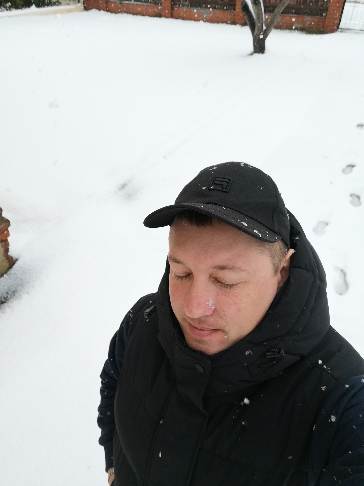 An overhead shot captures an individual standing outdoors amidst a fresh snowfall. The subject, an adult seen from the chest up, is wearing a black baseball cap with a dark, rectangular geometric logo on the front, and a black puffer jacket or vest over a dark long-sleeved garment. Their eyes are closed, and specks of snow are visible on their nose, the brim of their cap, and their shoulders, indicating active snowfall.
The scene is dominated by a blanket of pristine white snow covering the ground. Faint footprints can be seen in the snow on the right side of the frame, leading away from the subject. In the blurred background, a brick wall or fence runs horizontally, and the bare trunk of a tree stands further back. The light suggests it is daytime, likely overcast due to the snow. The overall impression is one of a quiet, cold winter day, with the person seemingly in a moment of calm, experiencing or enjoying the falling snow. No specific location identifiers or readable text are visible.