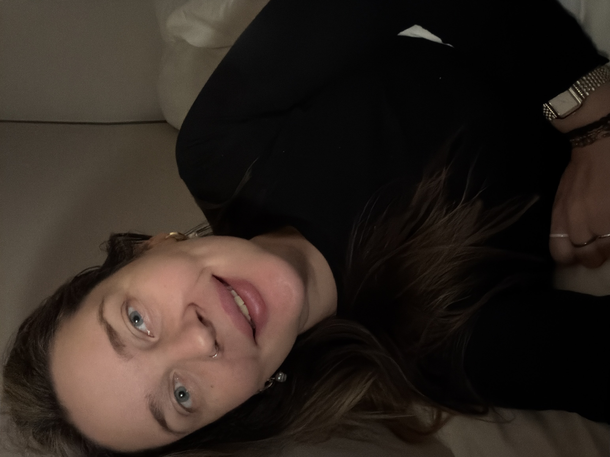 A young to middle-aged woman is depicted in a close-up, horizontal selfie. She is lying down, likely on a bed or sofa, with a soft, neutral-toned background suggesting indoor surroundings. The setting is in Moscow, Russia.
The woman has fair skin, blue eyes, and light brown hair that falls around her head. She is wearing a black long-sleeved top. Her left wrist features a square-faced, silver-toned watch with a metal link band, along with at least one bracelet. On her right hand, multiple rings are visible. She has a small silver hoop nose piercing and a visible silver-toned dangle earring on her left ear.
Her expression is relaxed and pleasant, with her lips slightly parted in what appears to be a soft smile or natural repose. Her gaze is directed straight at the camera. The lighting is dim and warm, indicating an indoor setting, possibly during the evening or night. No specific activity beyond posing for the camera is discernible. There is no visible text in the image.