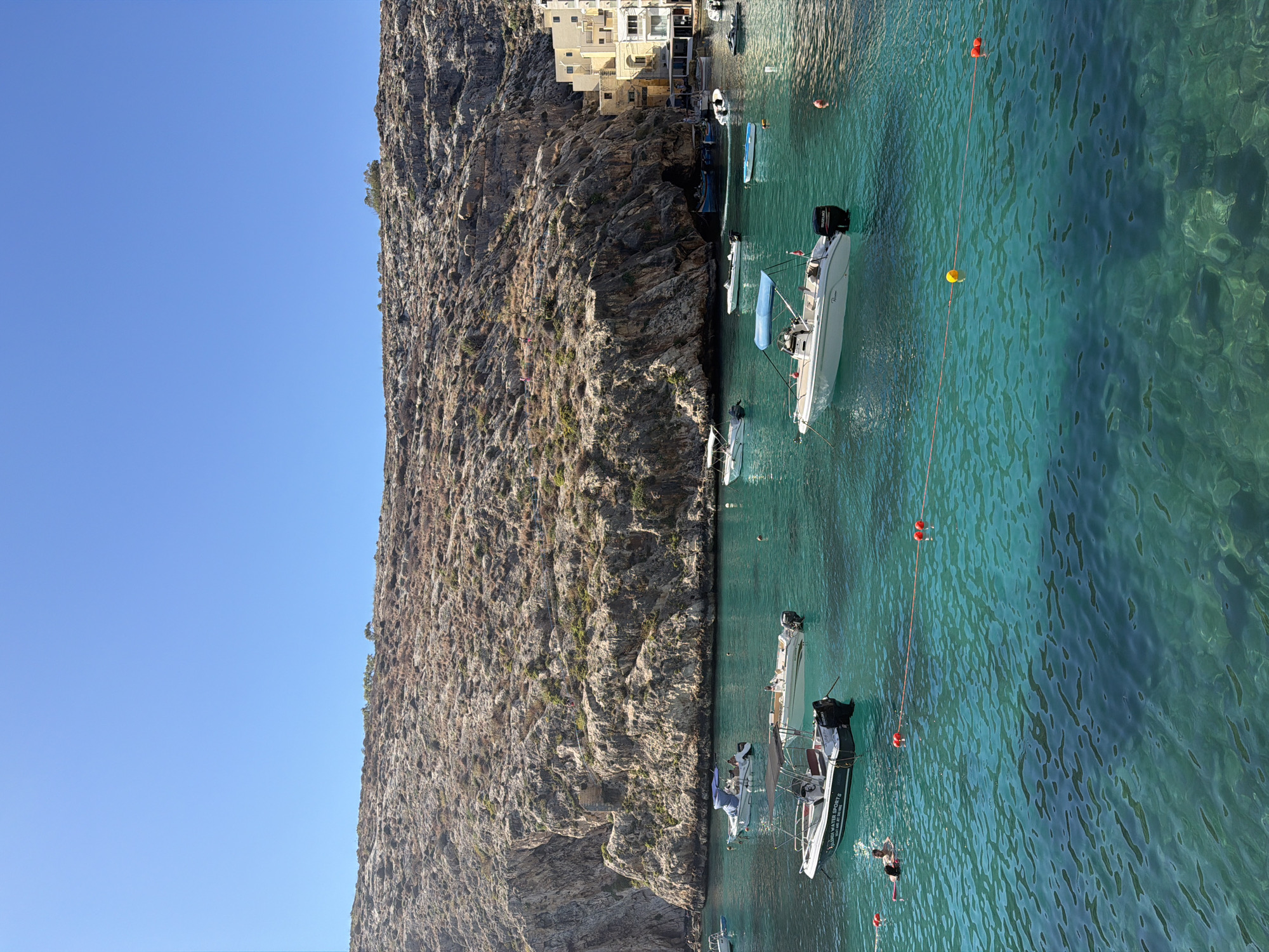 The image depicts a sunny day in Xlendi, Malta. The scene is dominated by a clear, turquoise body of water, likely Xlendi Bay, with several boats of various sizes moored within it. In the background, a rugged, rocky cliff face rises steeply, with a small cluster of yellow buildings visible perched near the water's edge on the right side of the frame. The water is calm and transparent, allowing the seabed and rocks to be seen in the shallower areas. One of the boats in the foreground displays the text "XLENDI WATER SPORT". Several people are visible in the water, some swimming and others near boats. The sky above is a vibrant, cloudless blue.
