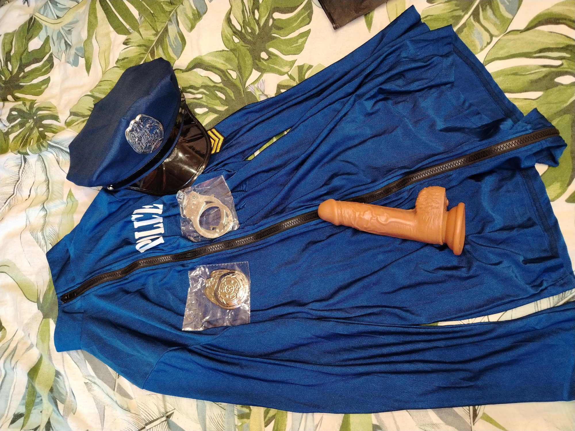 A blue police uniform lies on a bed with a patterned botanical print bedspread. The uniform consists of a zippered jacket with the word "POLICE" printed across the chest and matching trousers. A police cap rests on the left side of the uniform. A pair of handcuffs in clear plastic packaging and a police badge in a clear plastic bag are also on the uniform. A brown adult sex toy is positioned on the lower right portion of the uniform. There is no indication of the location of the photo as Kuyeda, Russia.