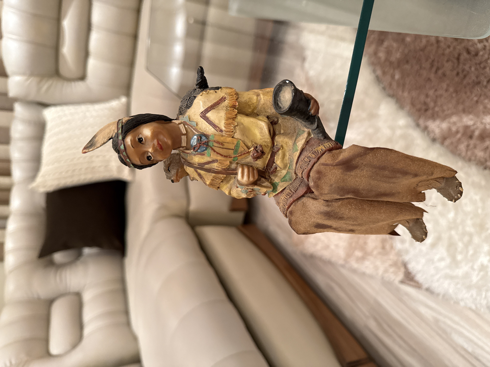 A detailed, close-up, eye-level shot captures a decorative figurine of a Native American child seated on a glass surface. The figurine, made of resin, depicts a young child with dark hair adorned with a feather and a beaded headband. They wear a yellow tunic with fringe detailing and brown trousers. The child holds a dark horn-shaped object in their right hand and has a small animal figure perched on their left shoulder. In the background, a large, plush, cream-colored sectional sofa with tufted cushions and dark brown accent pillows is visible. The flooring appears to be a combination of light wood planks and a plush cream and brown rug. The scene is lit by soft, ambient lighting. There are no identifiable landmarks or contextual clues in the image that would suggest its location in Targovishte, Bulgaria.