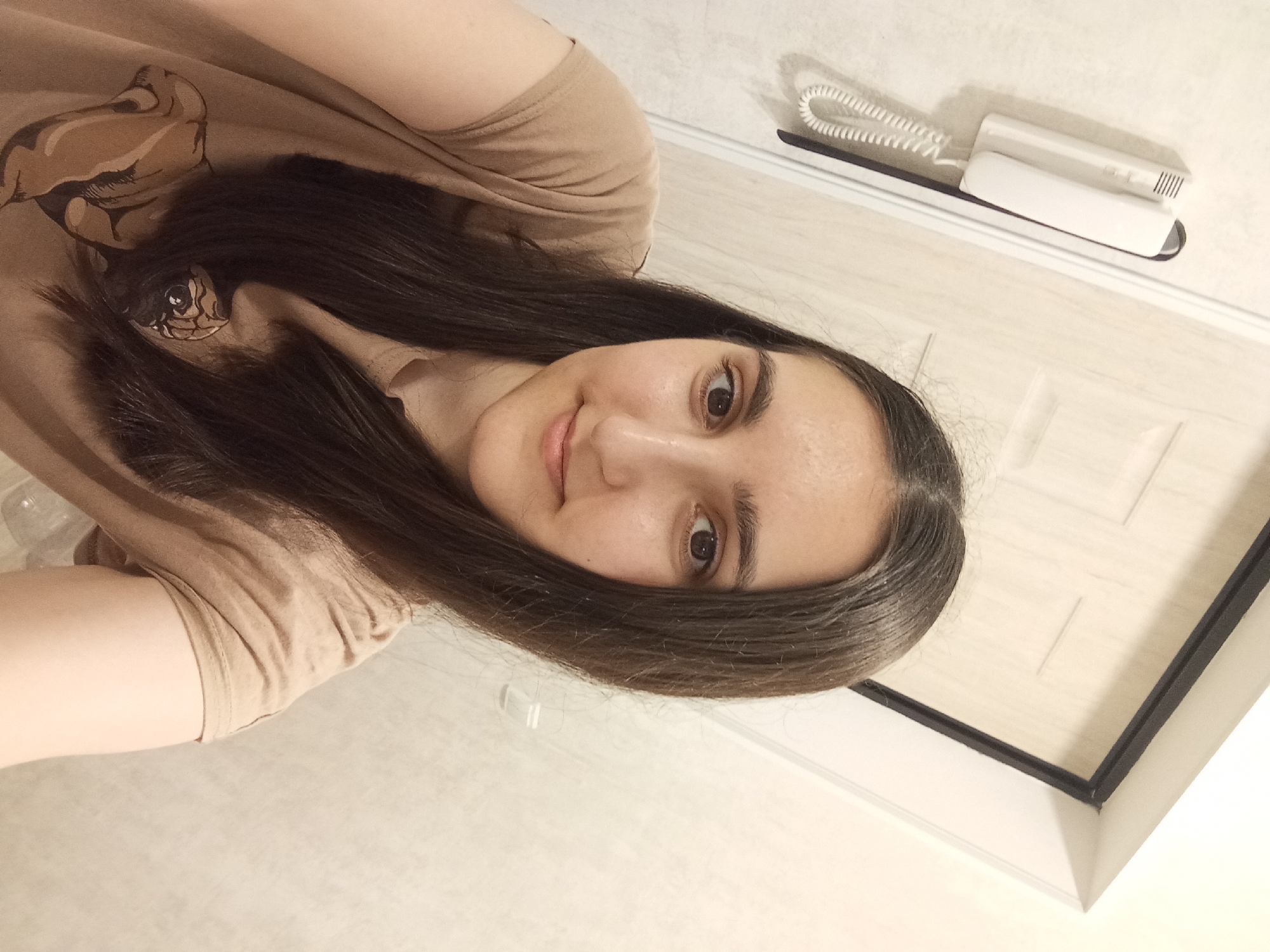 In an indoor setting, a young woman with long, dark hair takes a selfie. She is wearing a light brown t-shirt with a graphic print of a pug on the front. Her head is tilted slightly to the right, and she is looking towards the camera with a gentle smile. Behind her, a white door with vertical paneling is visible, and to her left, a white intercom unit is mounted on the wall. The lighting appears to be from an overhead source, casting soft shadows. The overall environment suggests a residential interior.
