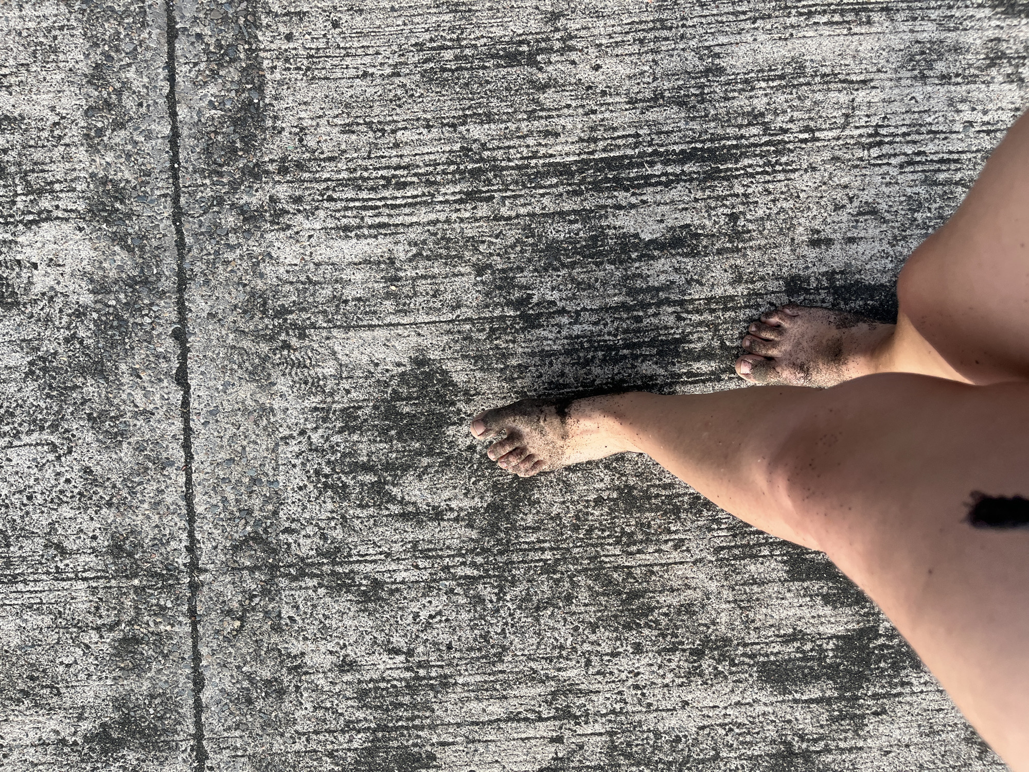 A high-angle, close-up shot shows a person's bare legs and feet walking on a concrete surface. The concrete is grey and textured with visible aggregate and parallel lines, suggesting it is a sidewalk or pathway. The person's feet and lower legs are partially covered in dark sand, indicating they have recently been at a beach. The shadow of the person falls on the concrete to the left of their legs. The scene is consistent with being in Ureki, Georgia, a location known for its magnetic black sand beaches and boardwalks.