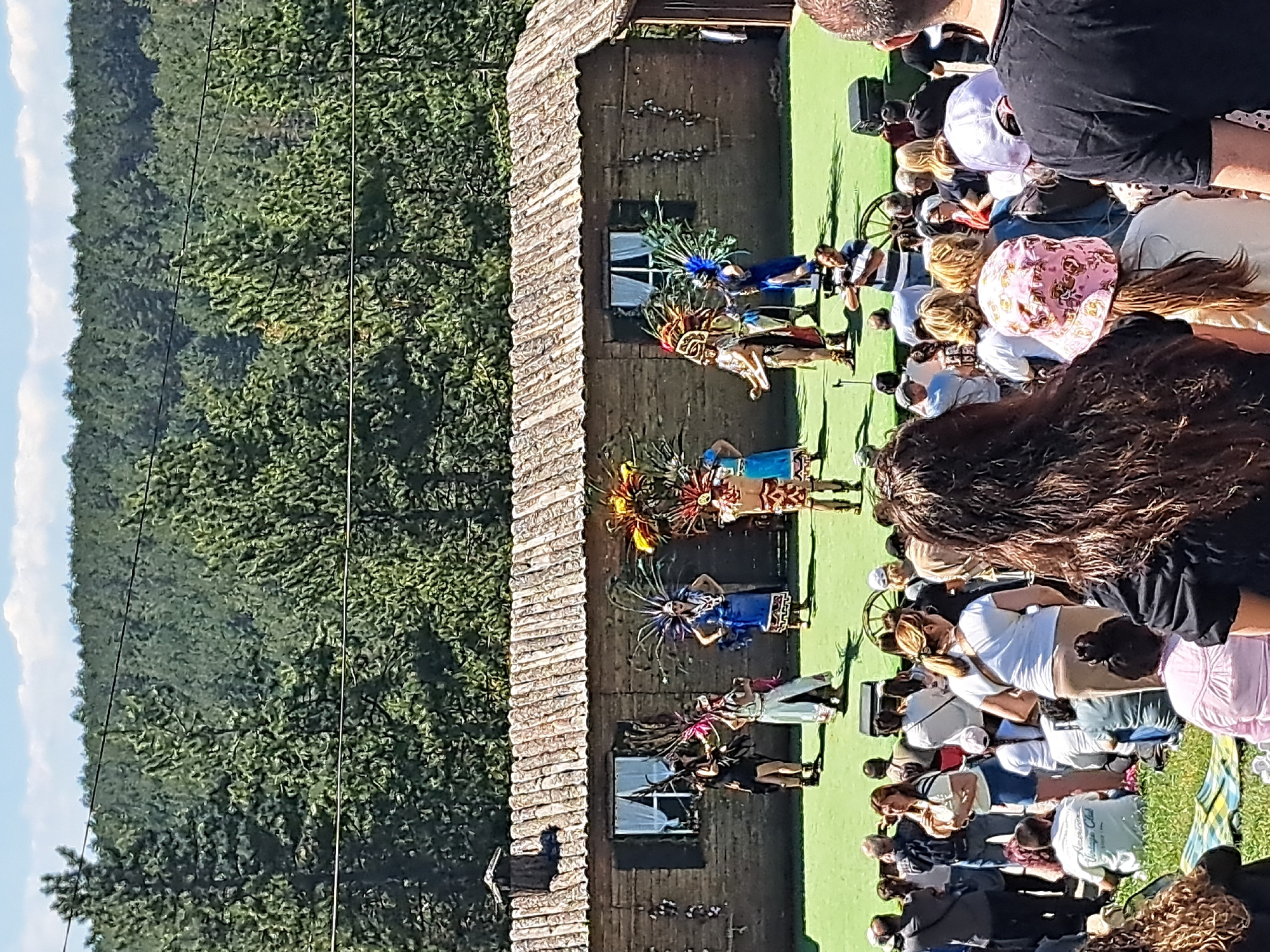 The image shows a group of individuals performing in what appears to be a traditional dance in front of an audience. The performers are wearing vibrant and colorful costumes, complete with headdresses. The performance is taking place outdoors on a green surface, possibly artificial grass. A rustic wooden building stands behind the performers, with small windows visible along the wall. In the background, a lush forest landscape is visible. The location appears to be in a rural or semi-rural area, in the city of Unknown, Unknown. The audience is composed of people of various ages and ethnicities, seated or standing and watching the performance. The overall scene conveys a sense of cultural celebration and community engagement.