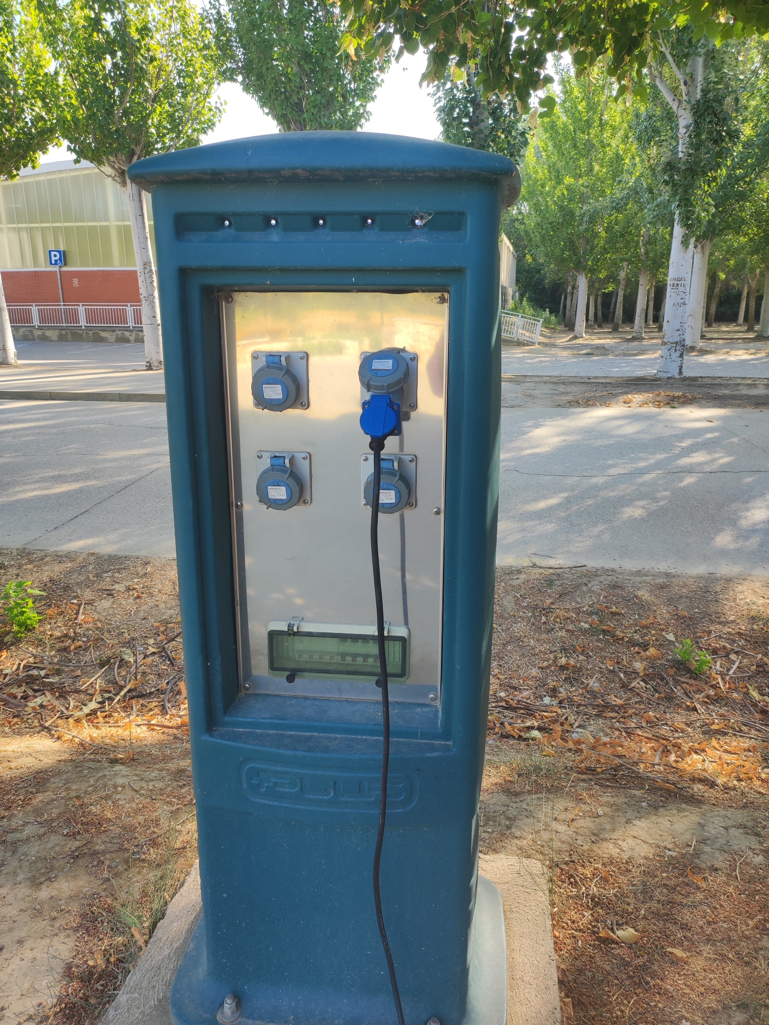 A teal colored electrical charging station stands on a concrete base on a paved walkway. The station has a silver panel with four electrical outlets, three of which are gray and one is blue. A black cable with a blue plug extends from the blue outlet. Above the outlets is a digital display. A logo is embossed on the front of the station. The station is surrounded by dry leaves and grass. In the background, there is a street, a building with a parking sign, and several trees. The scene appears to be in Villamuriel de Cerrato, Spain.